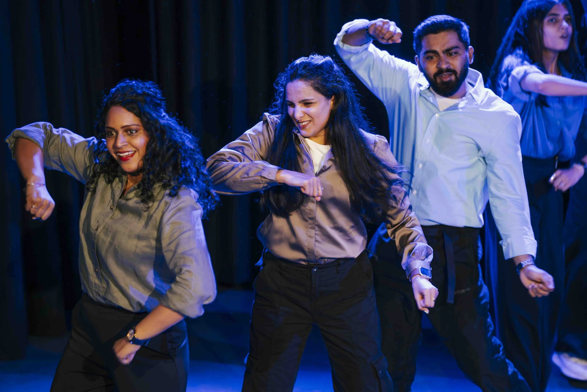 Group of people performing a dance on stage, with three women in front and two people in the background, all wearing casual clothes, under blue stage lighting.