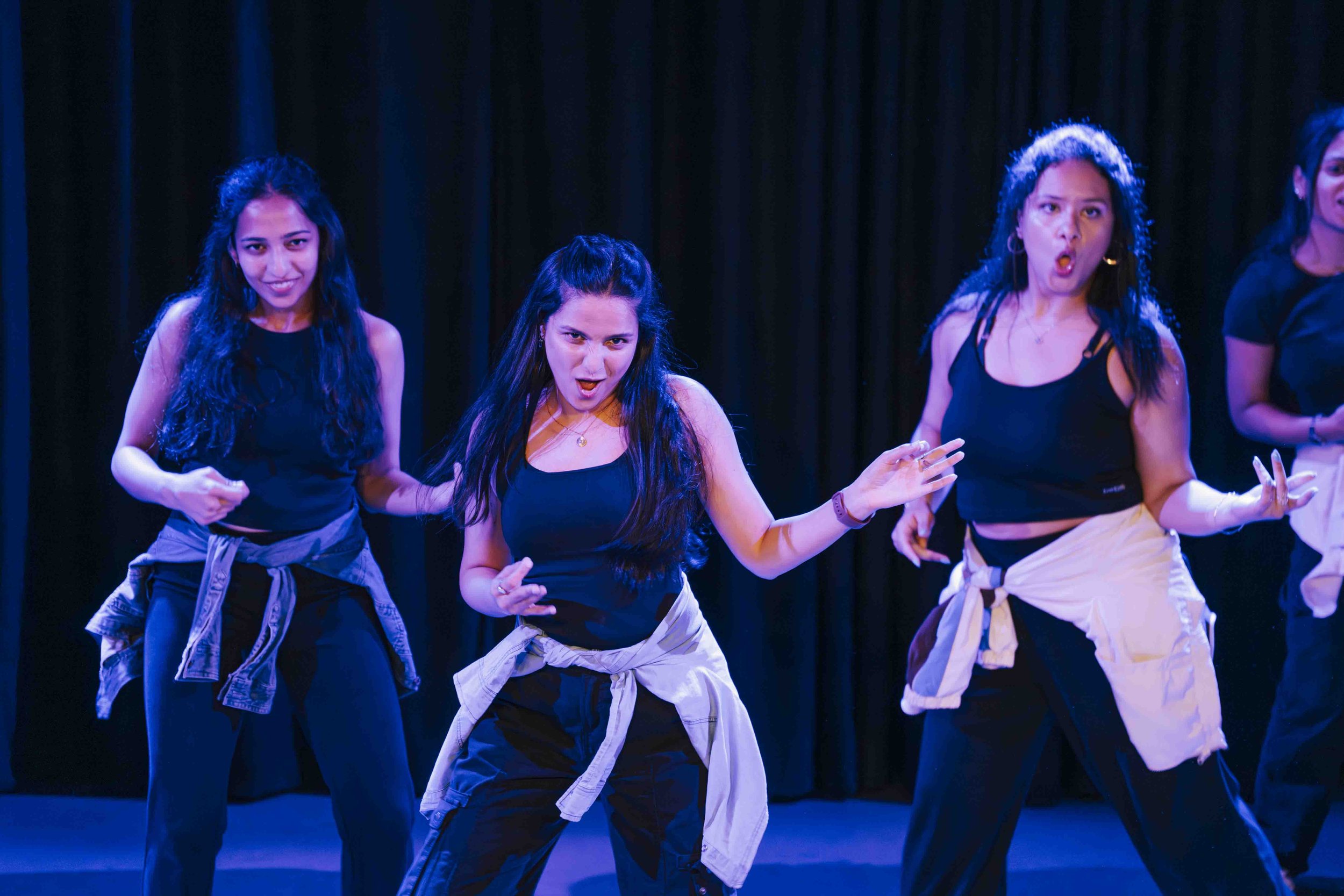 A group of women performing a dance routine on stage with a black curtain background, wearing black tops and pants, with some tied jackets around their waists.