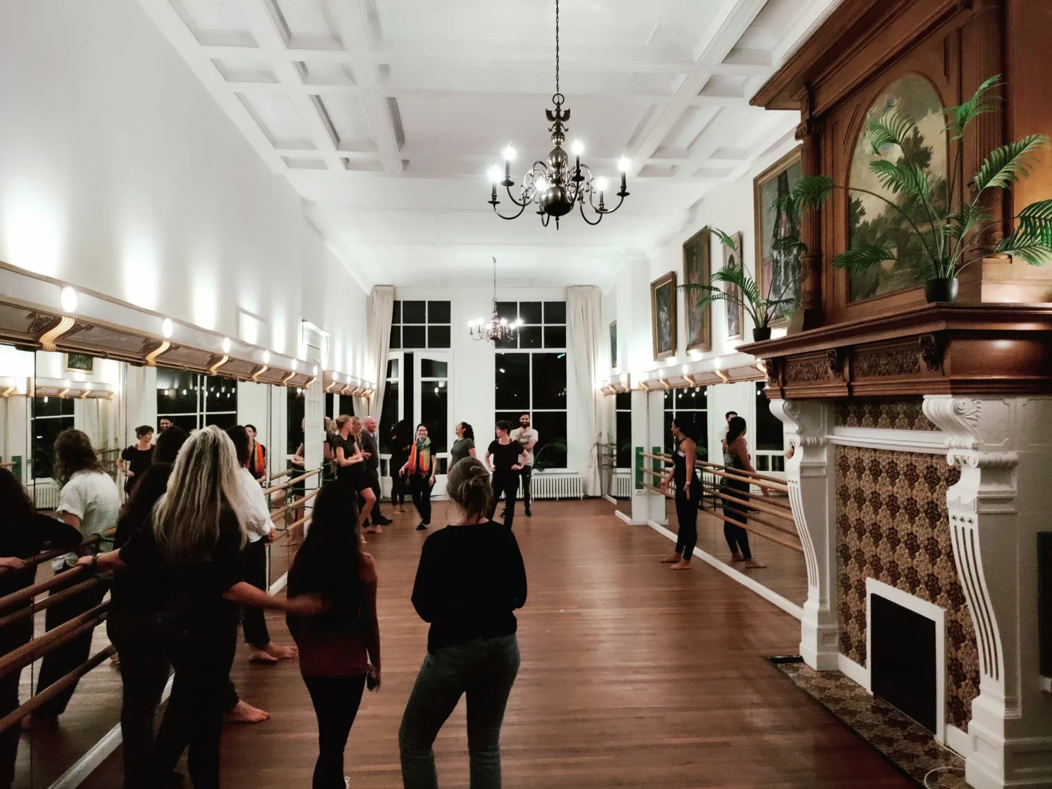 A group of people in a dance studio with mirrored walls and hardwood floors. The room has a large decorative fireplace, several potted plants, and chandeliers hanging from the ceiling. Participants are standing by ballet barre rails, engaged in a class or exercise.
