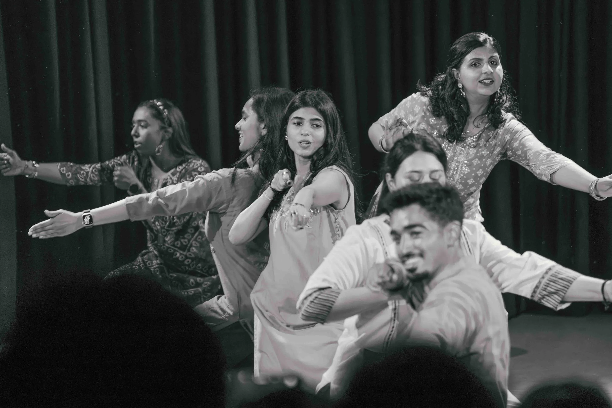 Group of people performing on stage with a dark curtain backdrop, mid-dance move, in black and white.