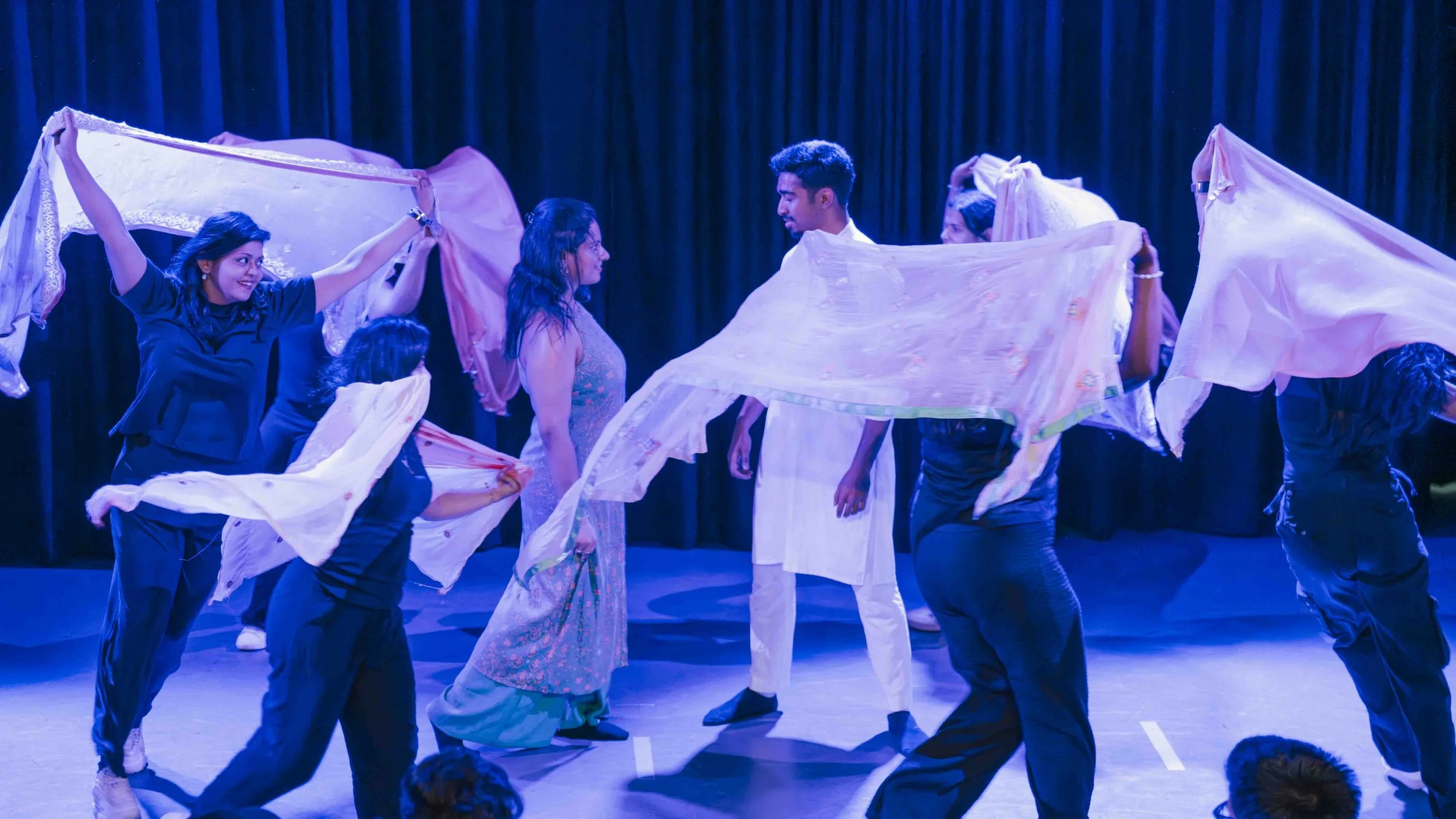 A group of people performing a dance or theatrical act, surrounded by pink and white cloths, with a dark stage background and blue lighting.
