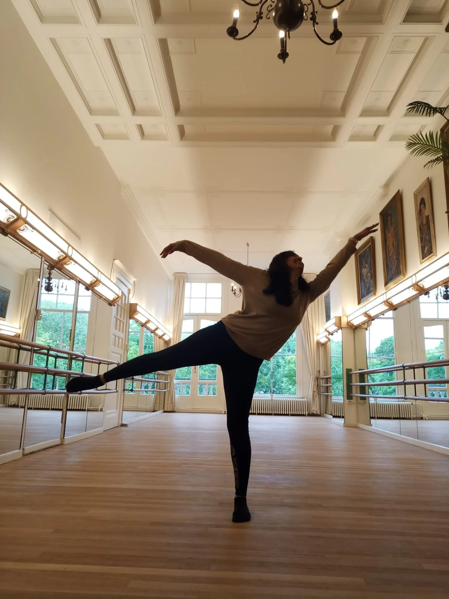 Person dancing in a ballet studio with wooden floors, mirrors, and a high ceiling.