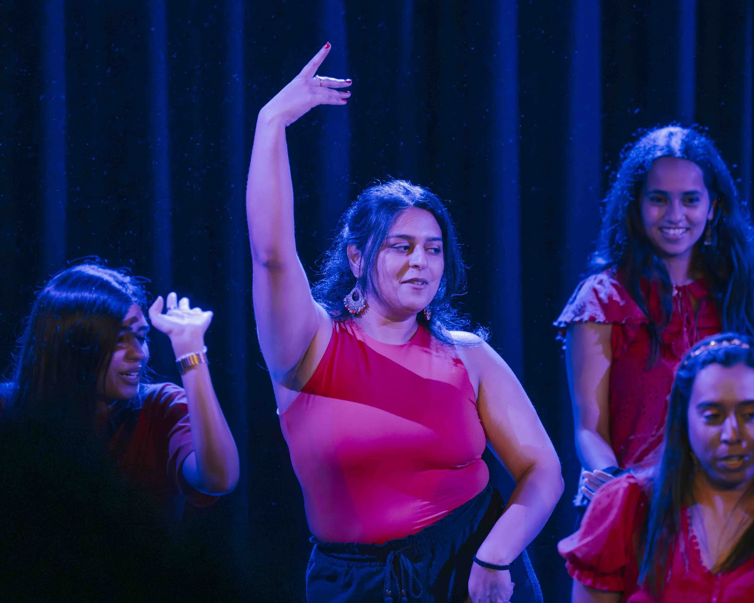 A woman in a red top and black pants dancing with her hand raised, surrounded by other women also dancing, in a scene with a dark curtain background.