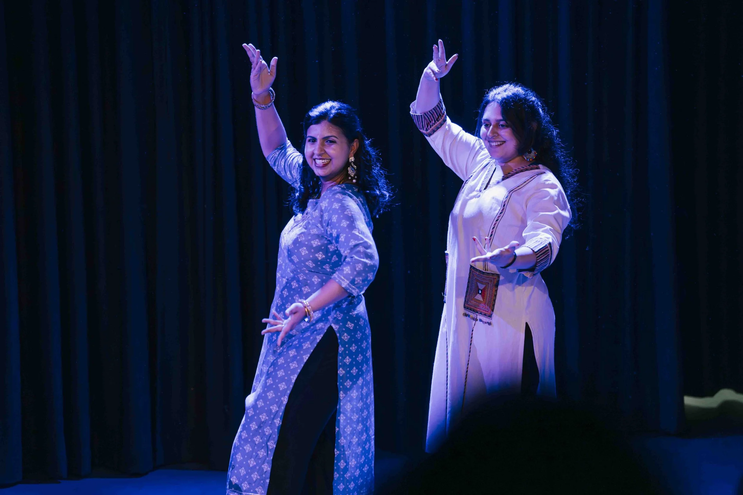 Two women dancing on stage with dark curtains in the background, wearing traditional Indian clothing and smiling.