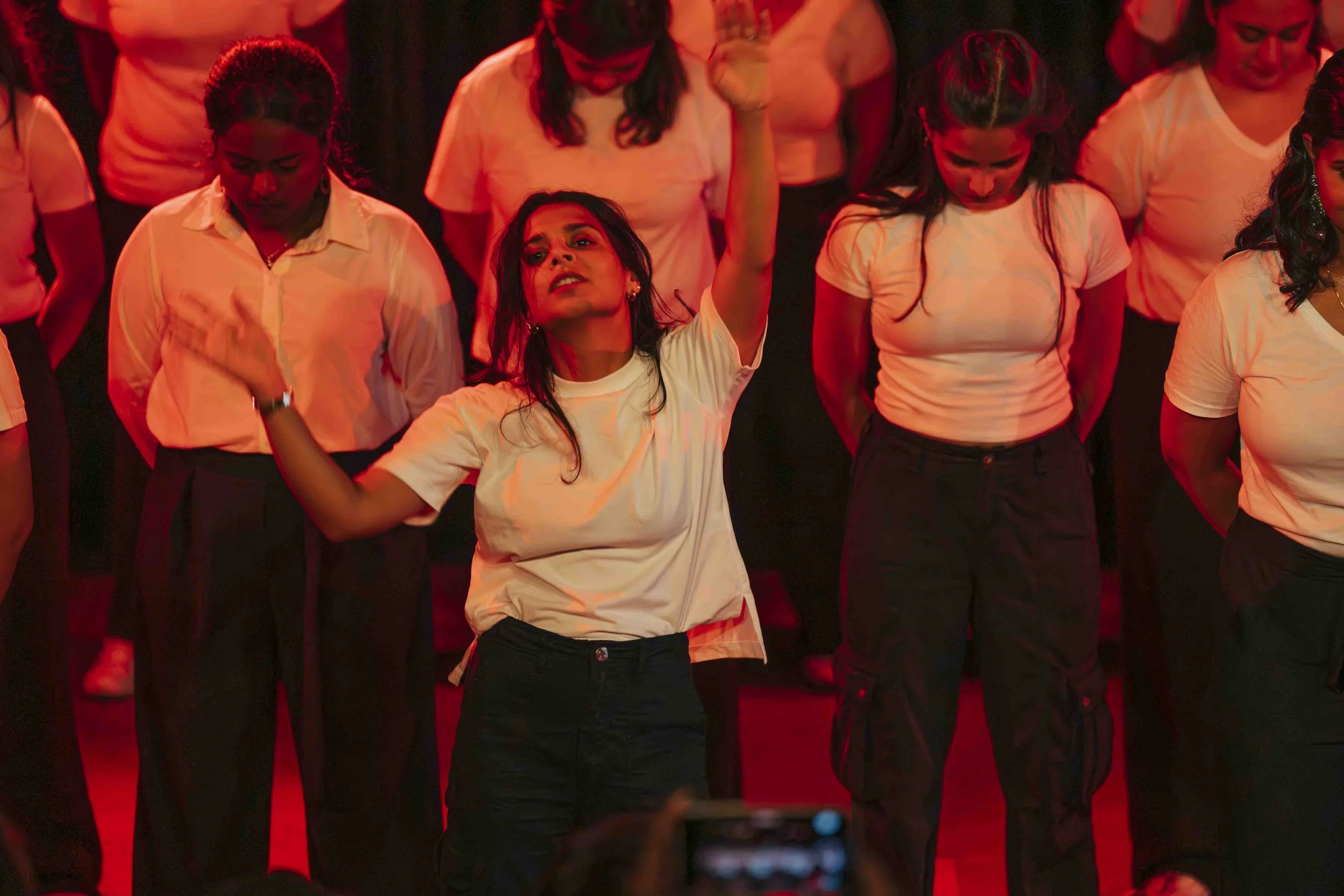 Group of women dancing on stage under red lighting, wearing white shirts and black pants.