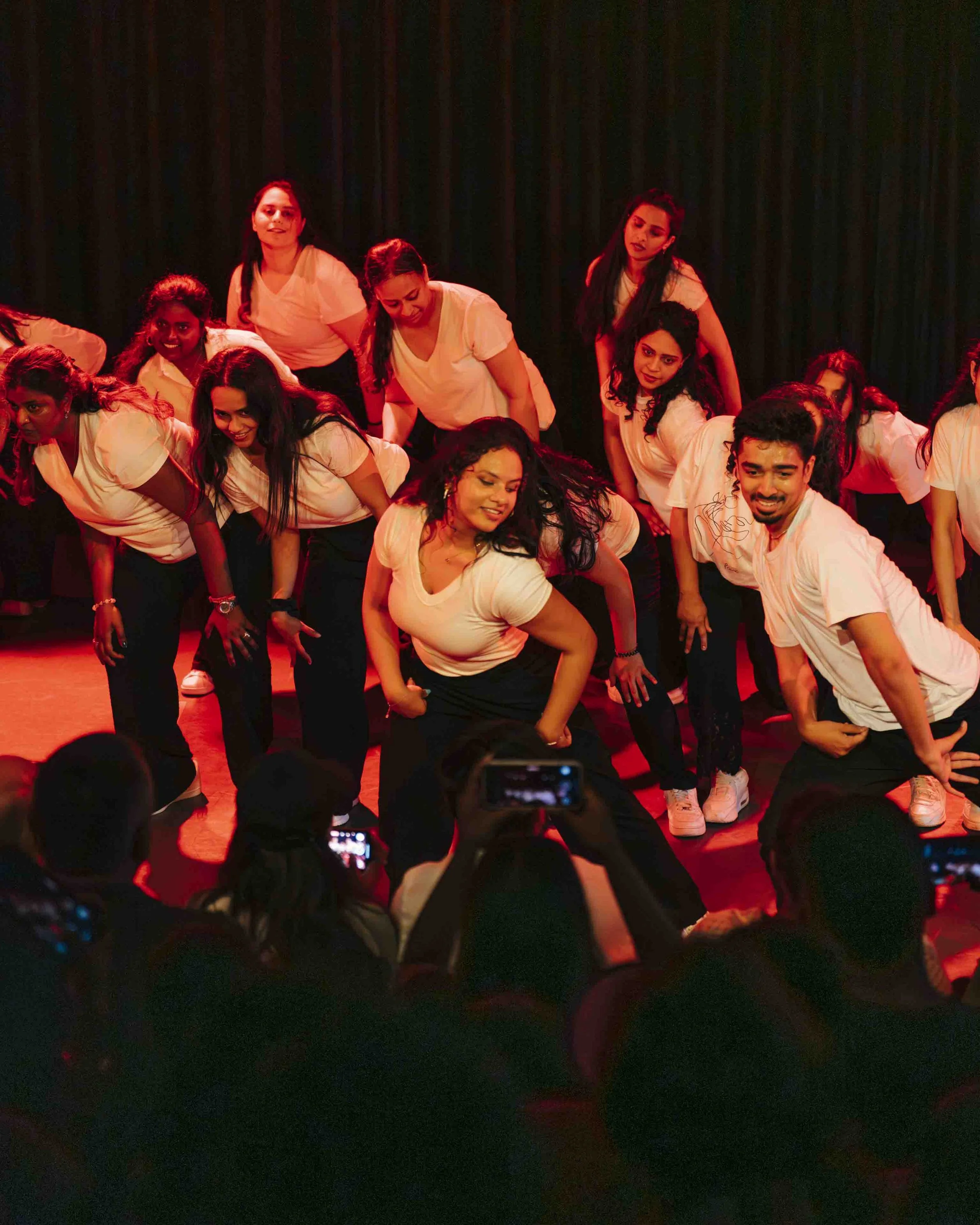 Group of people performing a dance on stage, with audience members taking photos in the foreground.
