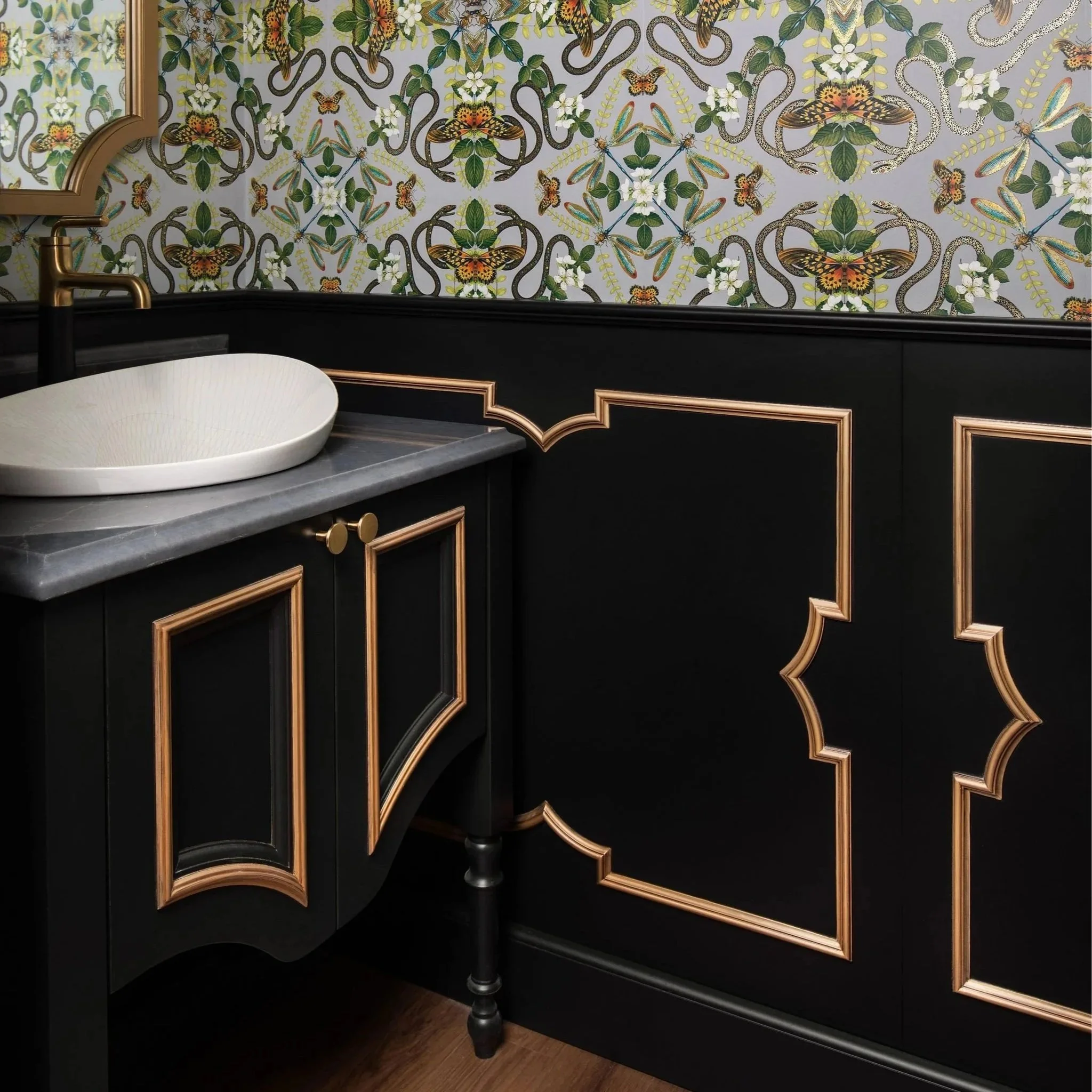 A bathroom with black cabinets featuring gold trim, a grey countertop, a white vessel sink, and patterned wallpaper with insects and floral motifs.