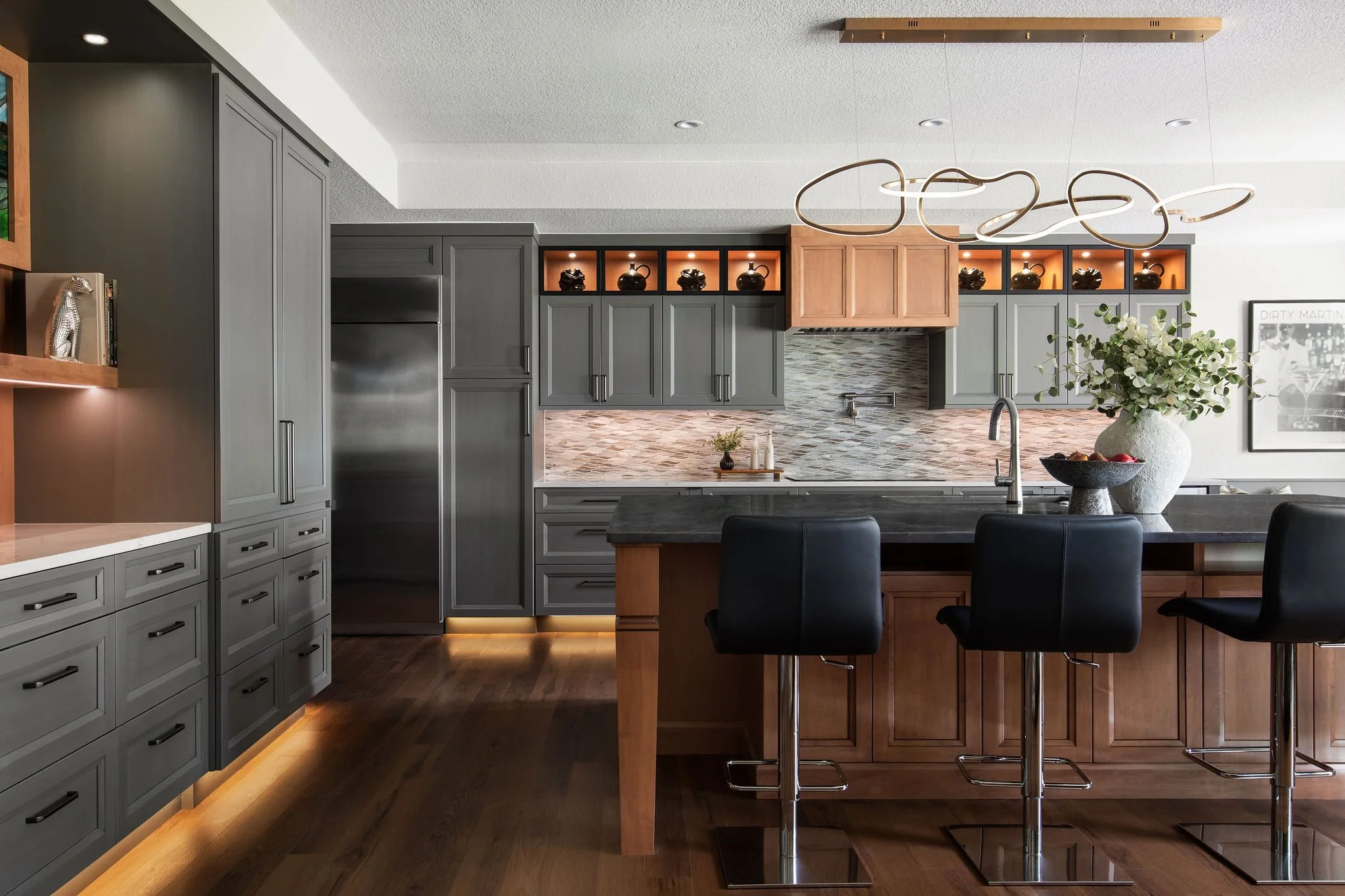 Modern kitchen with gray and wooden cabinets, black bar stools, and a dark countertop island. There are decorative lighting elements, plants, and framed artwork on the wall.