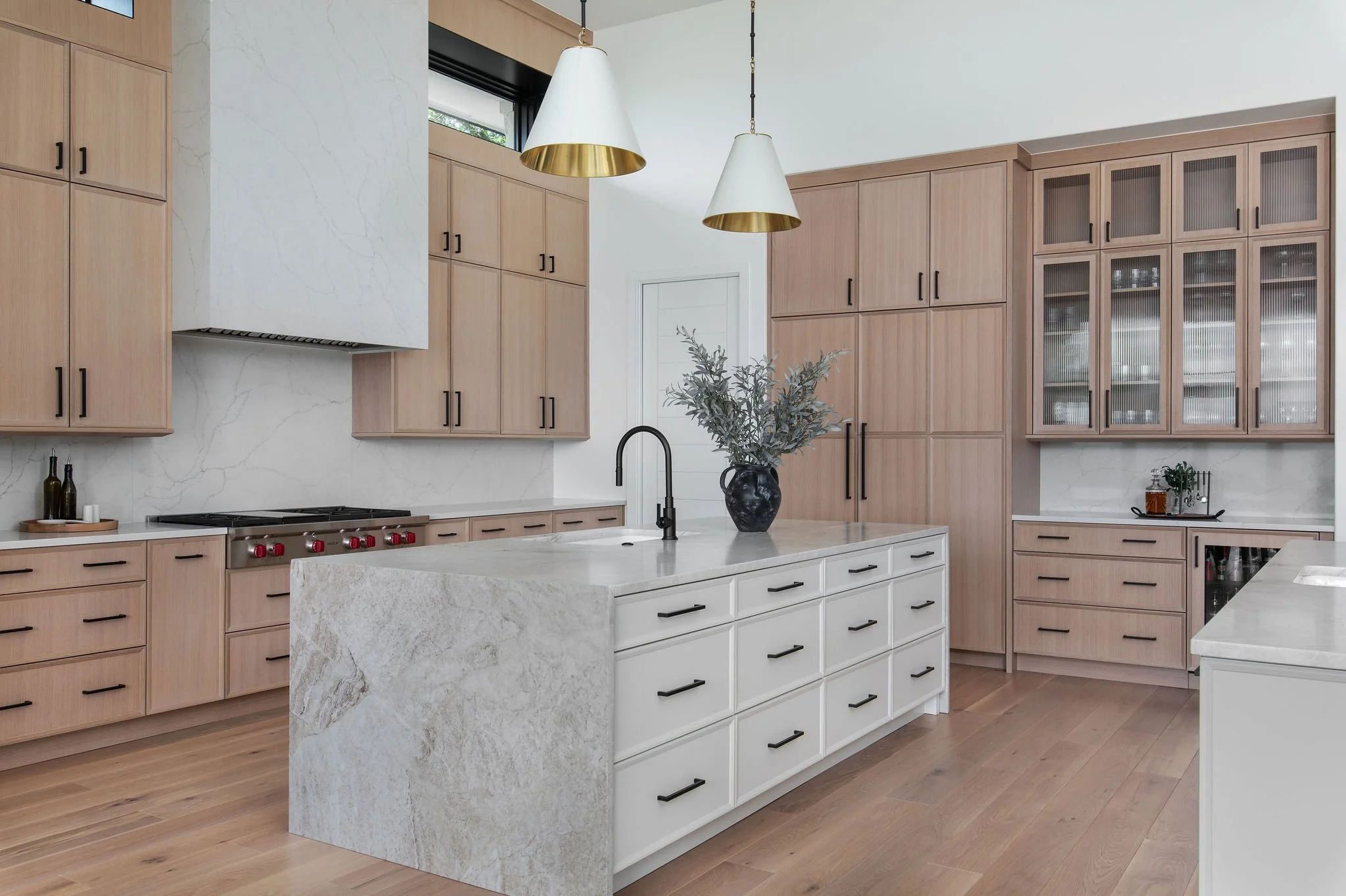 Modern kitchen with light oak wood cabinets, a large marble look quartzite island, black fixtures, pendant lights, and decorative plant vase.