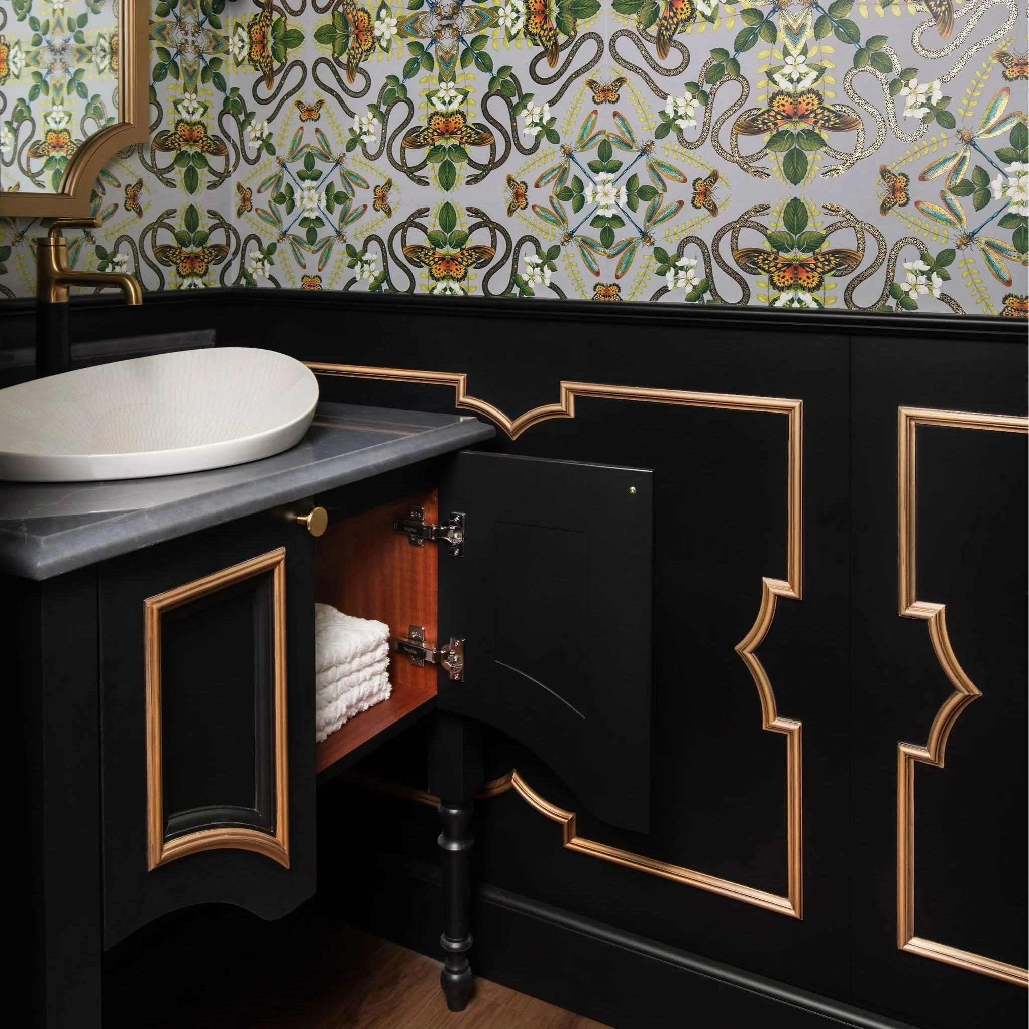 Modern kitchen with navy blue cabinets, white countertop, and gold hardware. Includes stainless steel sink, custom paper towel holder, wicker baskets, and dining area in the background.