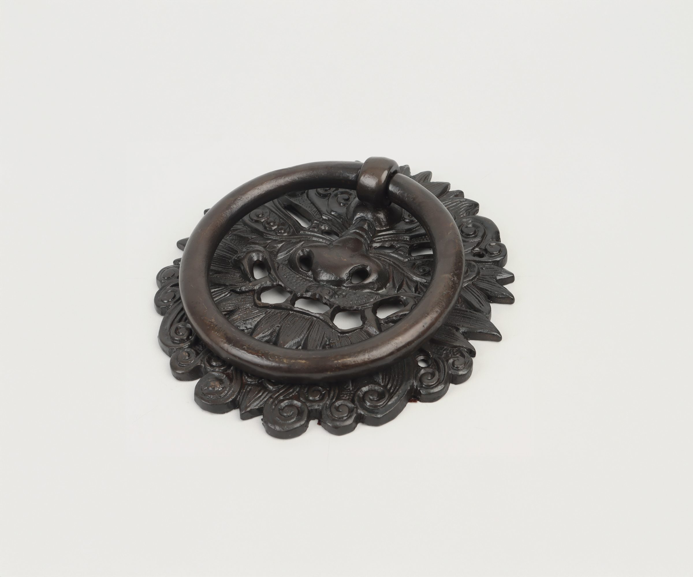 Decorative antique door knocker featuring a lion's face and a circular ring, mounted on an ornate metal backplate with floral and swirl patterns, set on a plain white background.
