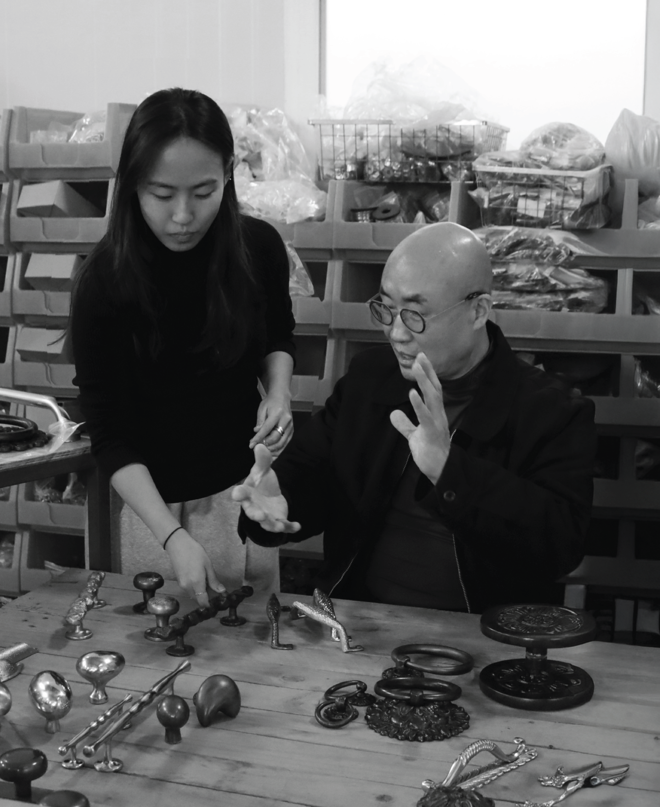 Two people, one woman and one man with glasses, are examining and discussing various Korean brass hardware pieces on a wooden table. The background shows shelves with boxes and plastic bags filled with more hardware items.