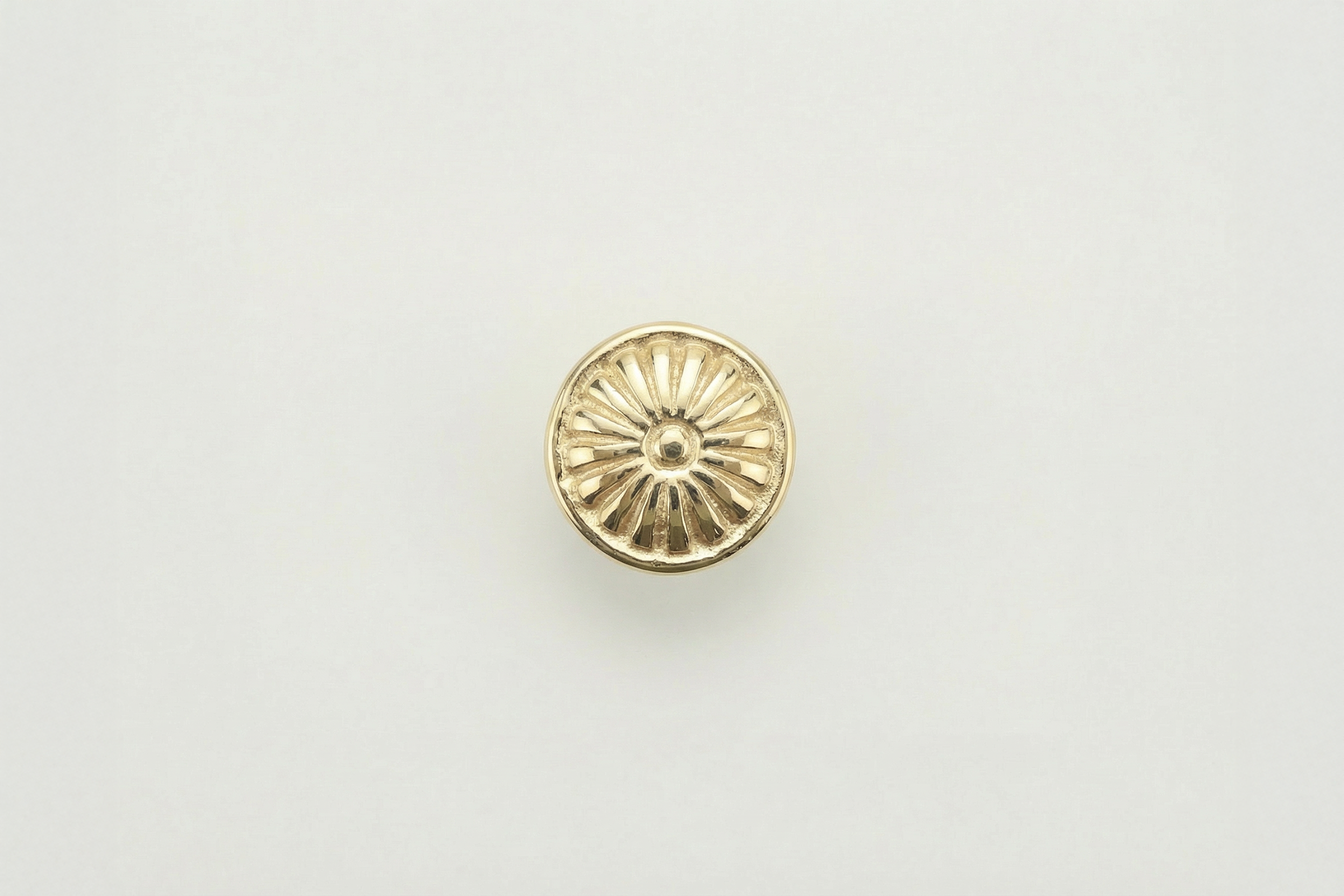 A small, circular, gold-colored object with a sunburst pattern, viewed from above on a plain white background.
