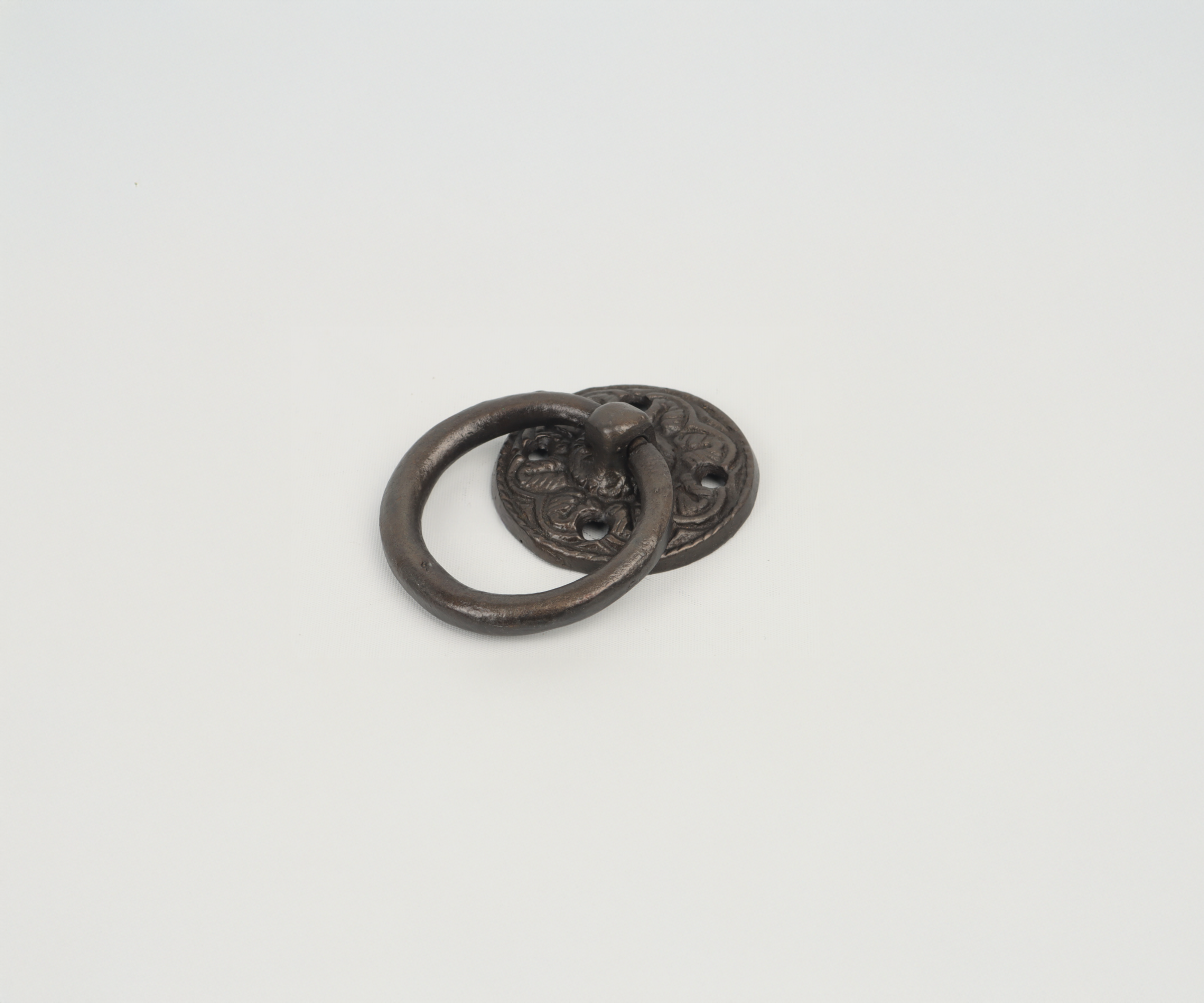 Antique metal door knocker with a circular ring attached to an ornate backplate, placed on a plain white surface.