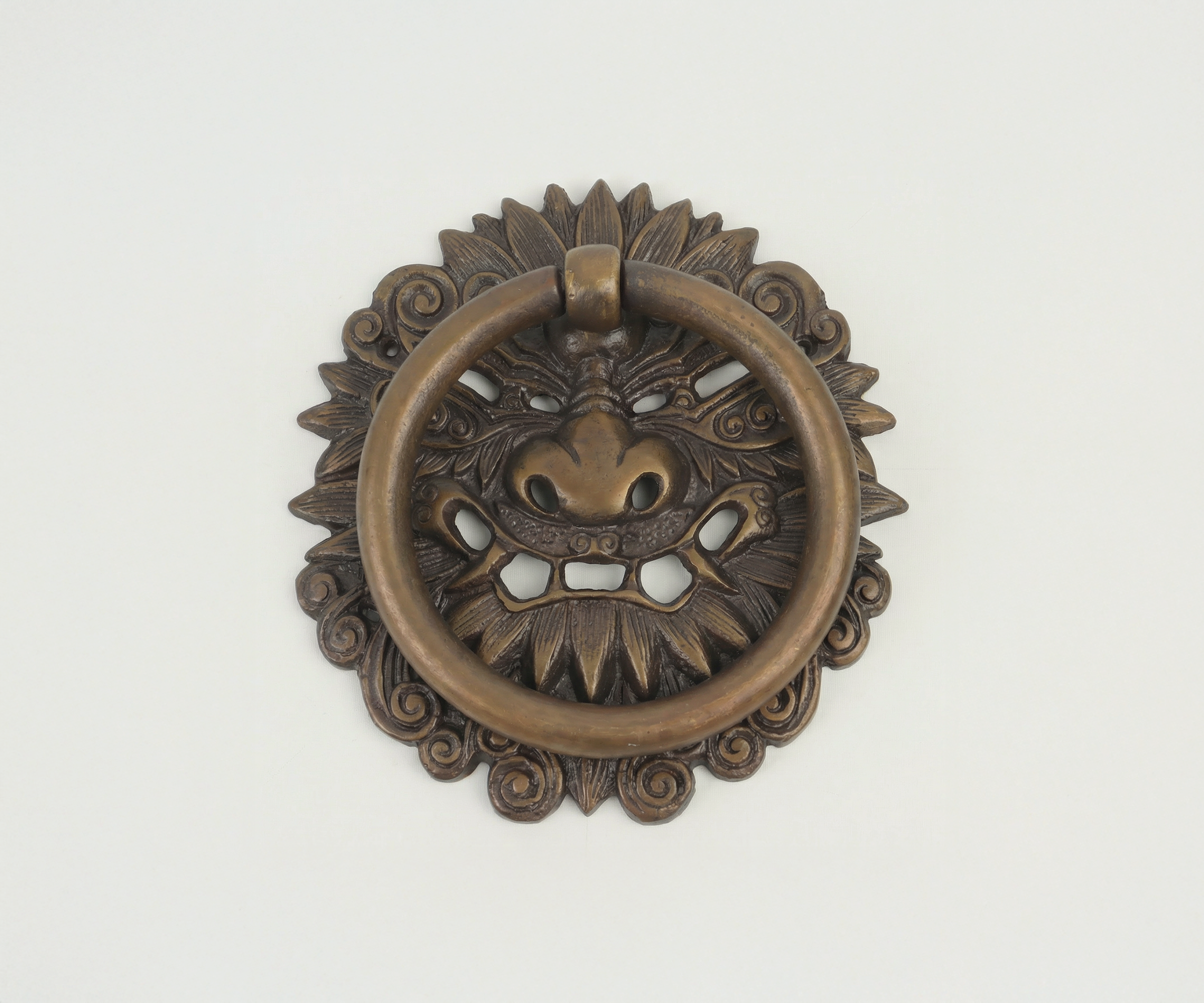 Bronze-colored Japanese-style lion face door knocker with a circular ring in the mouth, mounted on a plain white wall.