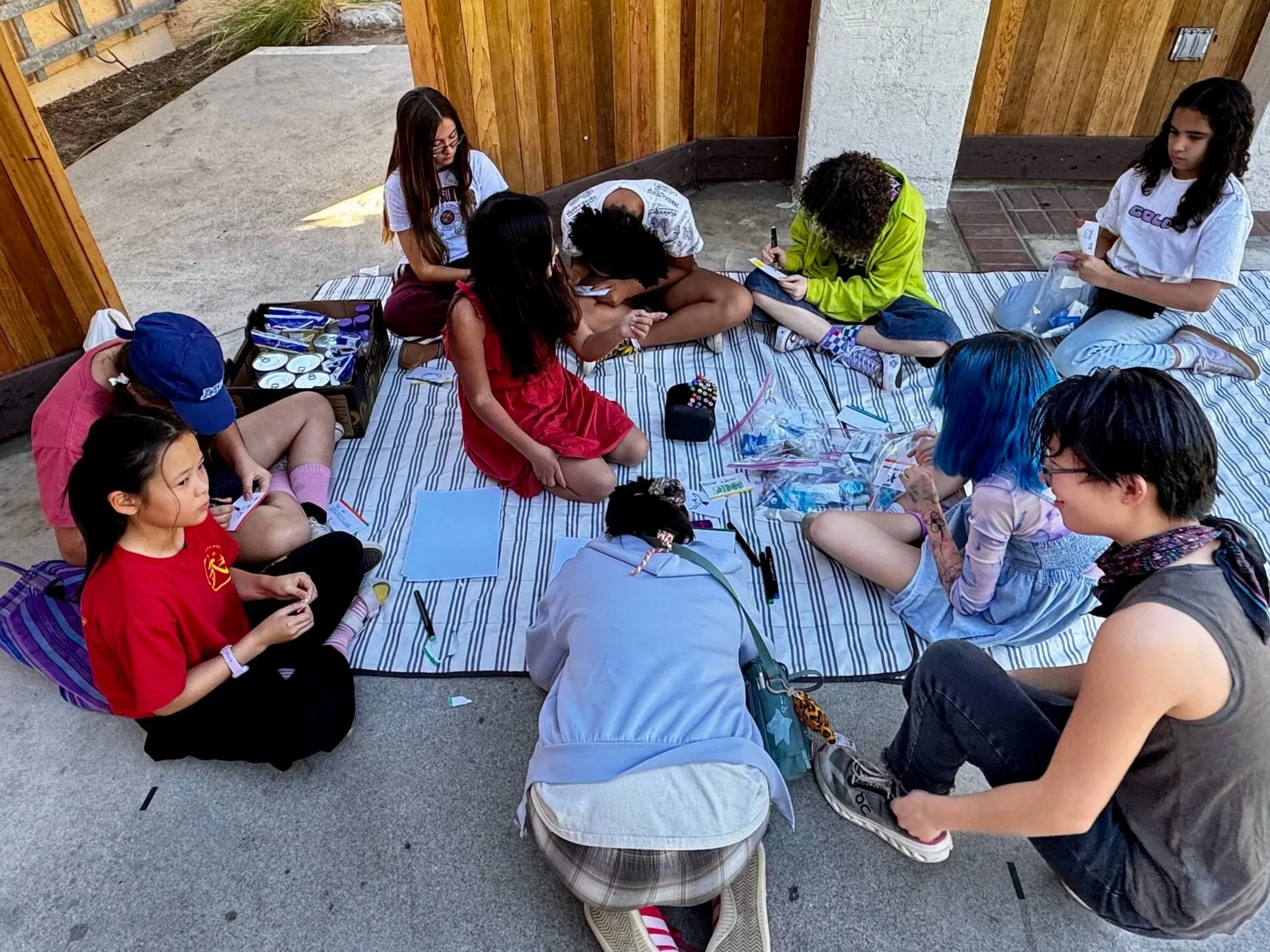 Group of young people volunteering working together to make kits for foster kids on a striped blanket, surrounded by art supplies, with wooden and concrete walls in the background.