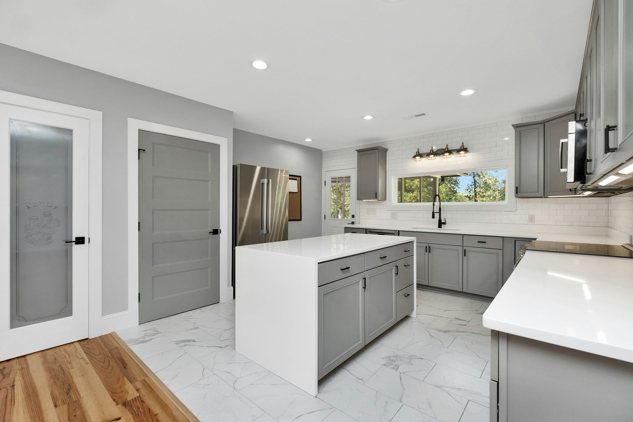 Modern kitchen with gray cabinets, white countertops, marble flooring, stainless steel refrigerator, black faucet, white subway tile backsplash, and a large window showing green trees outside.