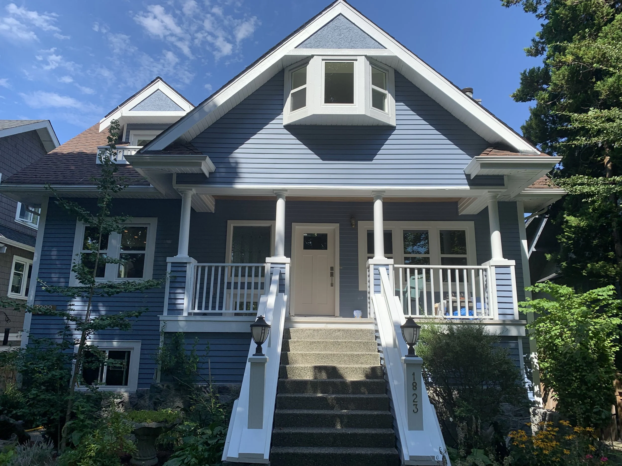Blue two-story house with white trim, front porch, and stairs leading up to the front door, surrounded by green trees and plants, under a blue sky.