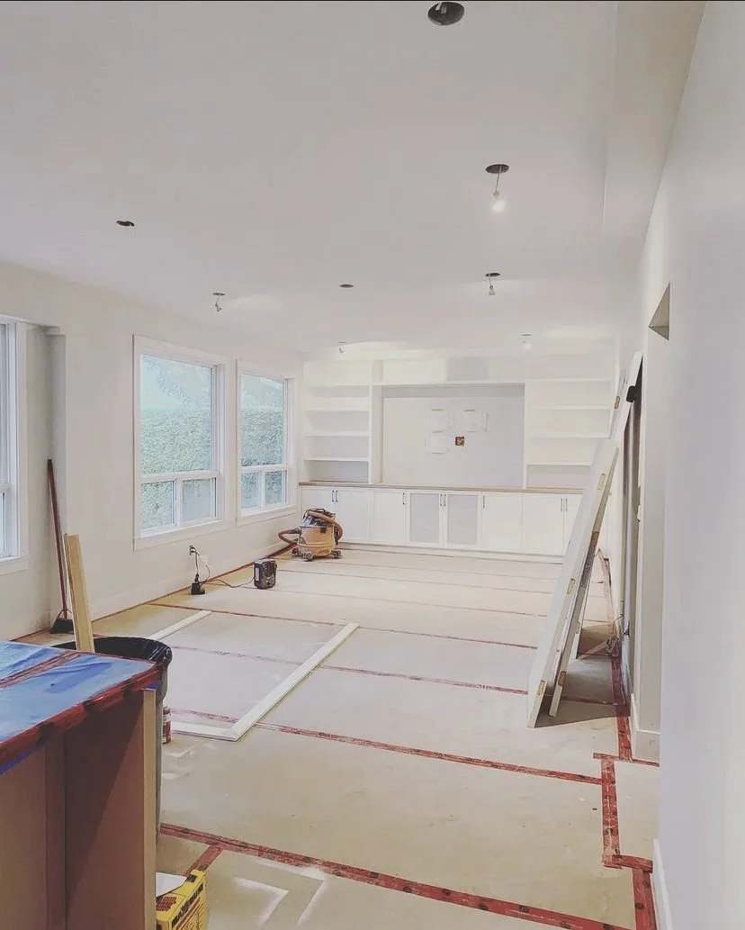 A room under construction or renovation with white walls and ceiling. The floor is unfinished with red tape markings, possibly for layout or safety. Several large windows allow natural light in. Cabinets are installed on the far wall, and there are construction tools like a ladder, a saw, and a bag on the floor, indicating ongoing work.