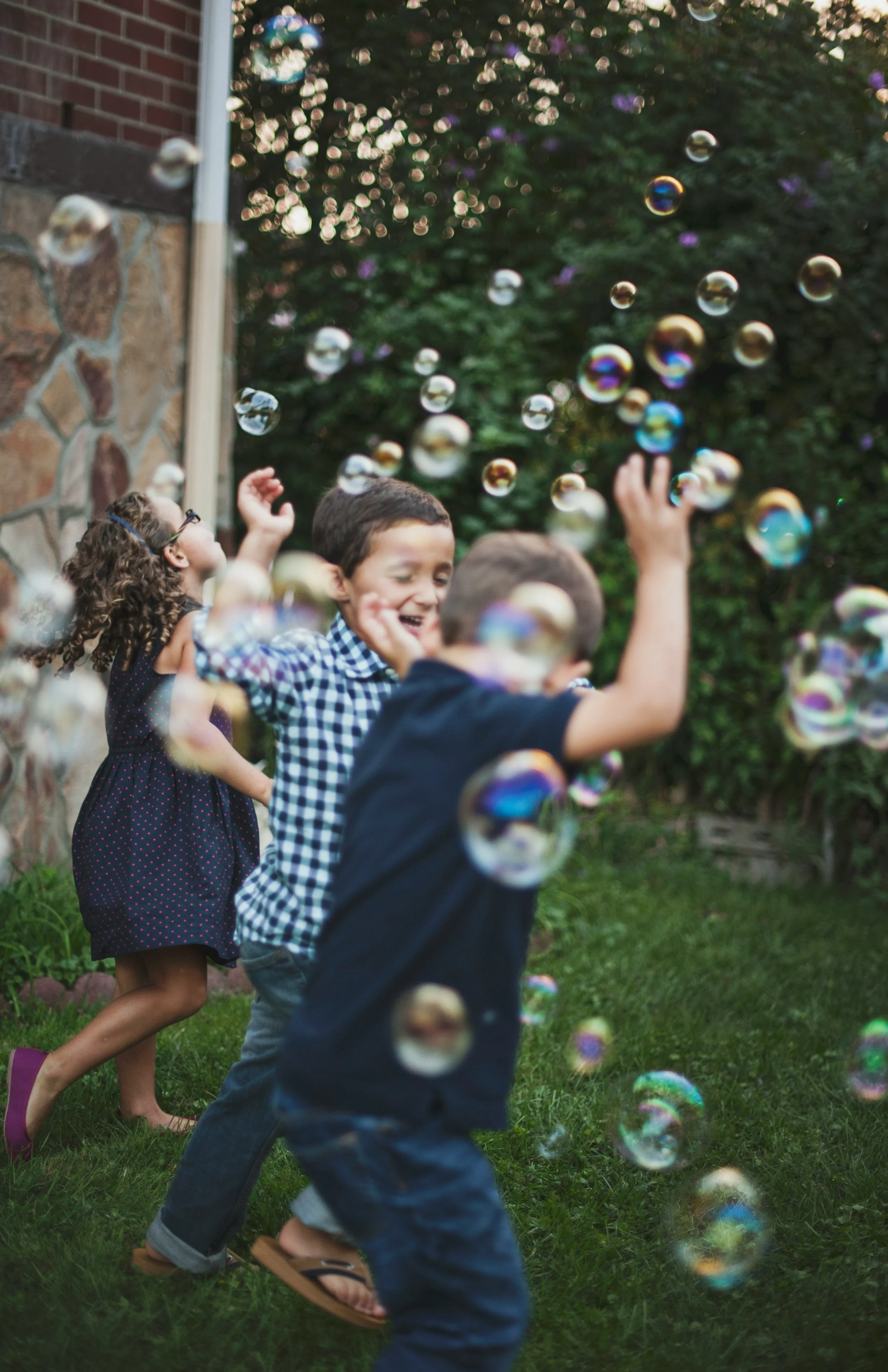 Children playing and laughing outdoors among floating soap bubbles on a grassy backyard with a brick wall and trees in the background.