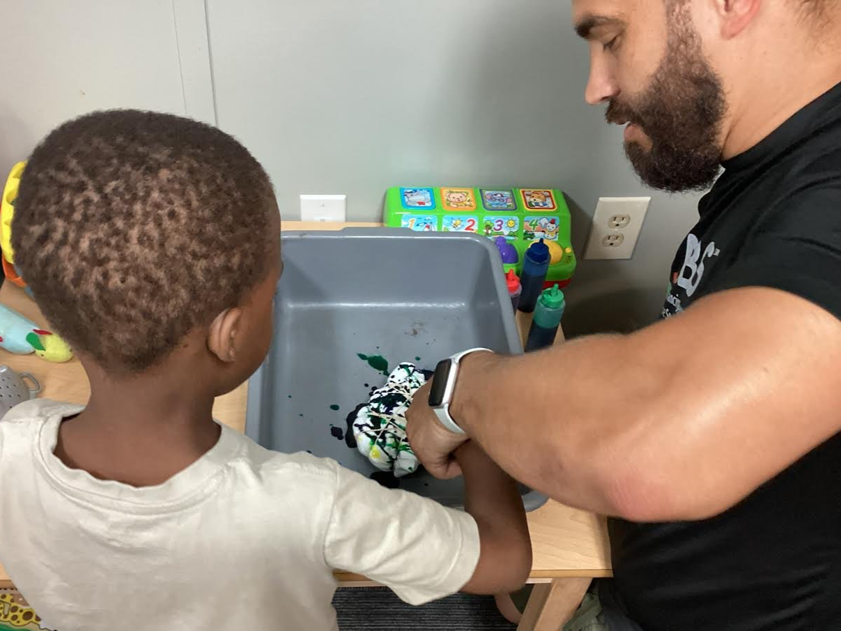 A man and a young boy washing a colorful tie-dye shirt in a plastic tub at a table.