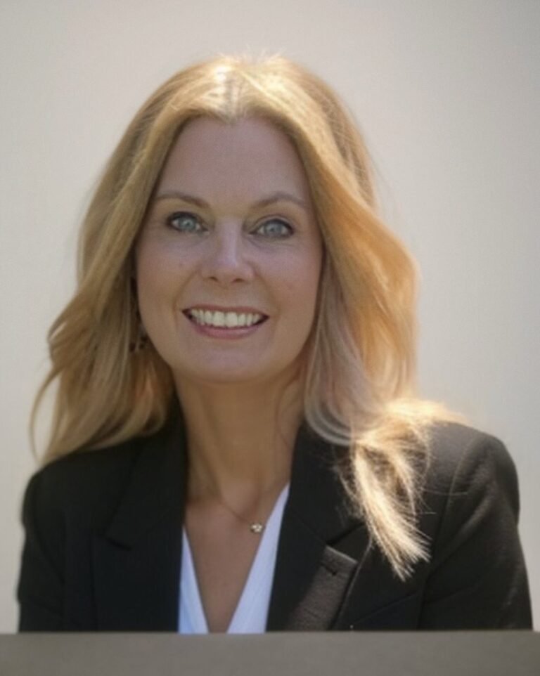 A smiling woman with blonde hair wearing a black blazer and white top, posing for a portrait against a plain background.
