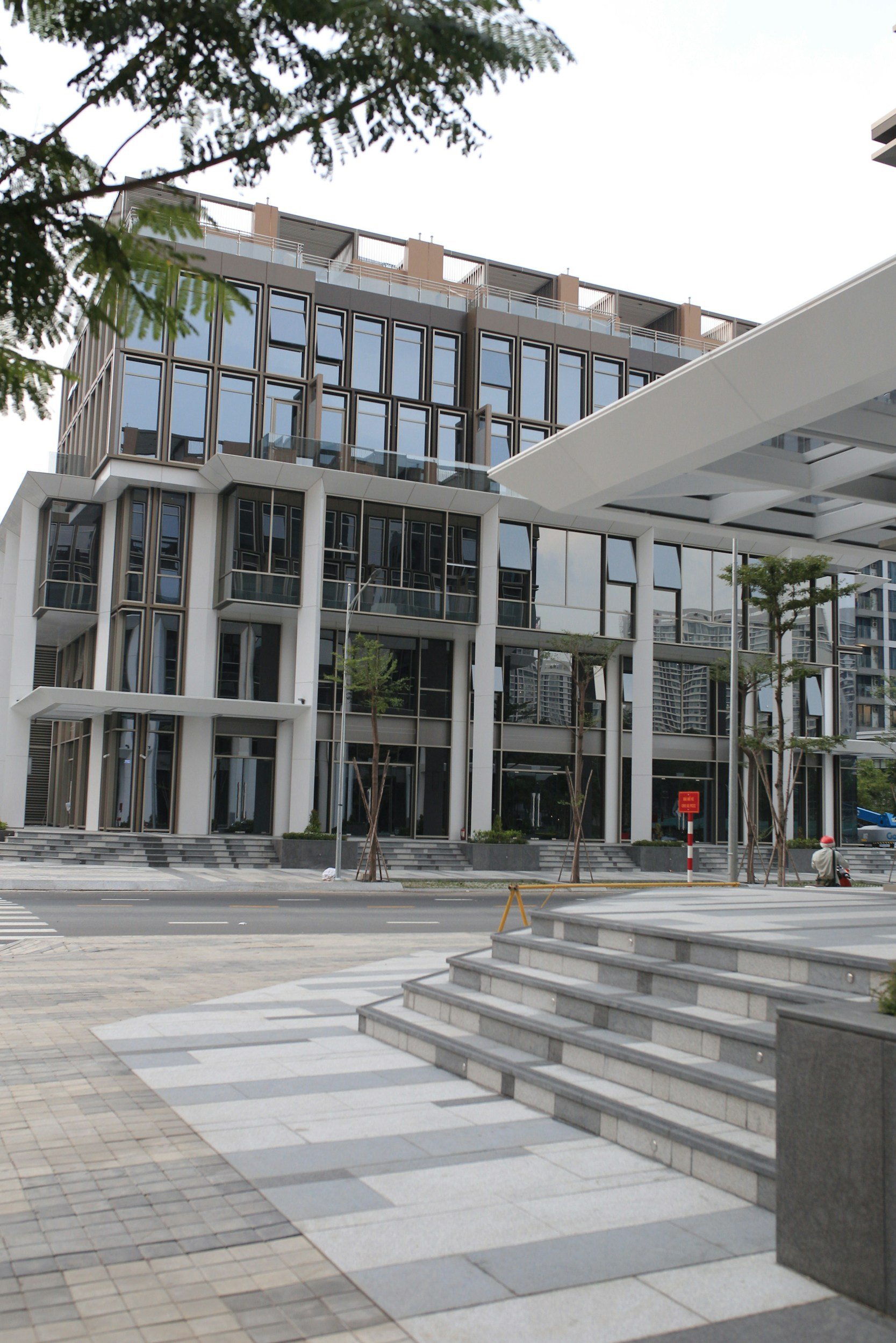 Modern multi-story building with glass windows and a white overhang, sidewalk with stairs, and trees in an urban setting.