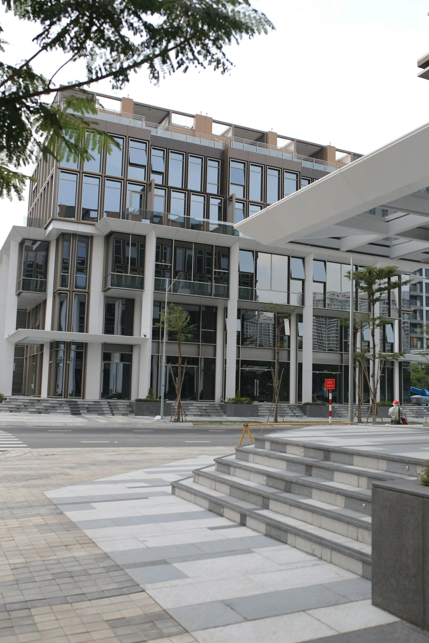 Modern multi-story office building with large glass windows, situated along a city street with steps and paved walkways in the foreground, trees, and a pedestrian in a helmet.