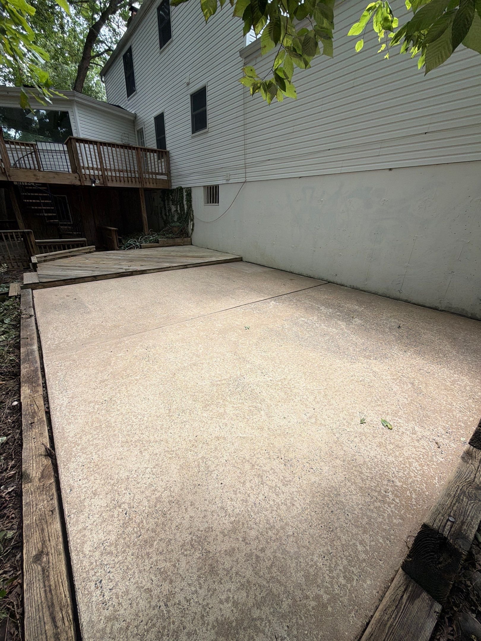 A concrete backyard patio area next to a house with white siding, a wooden deck, and a small wooden ramp. There are trees overhead and some garden hoses and plants along the border.