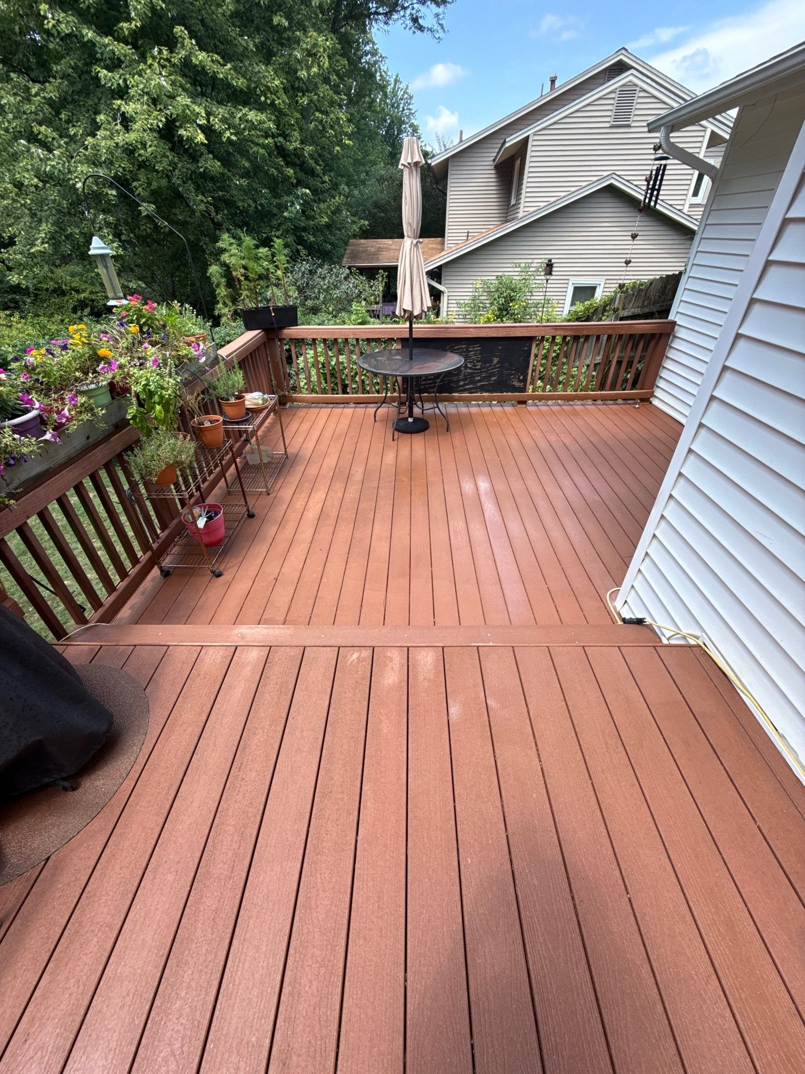 A wooden outdoor deck with potted plants, flowers, a small table with a closed umbrella, and a black object in the corner, surrounded by green trees and neighboring houses under a partly cloudy sky.