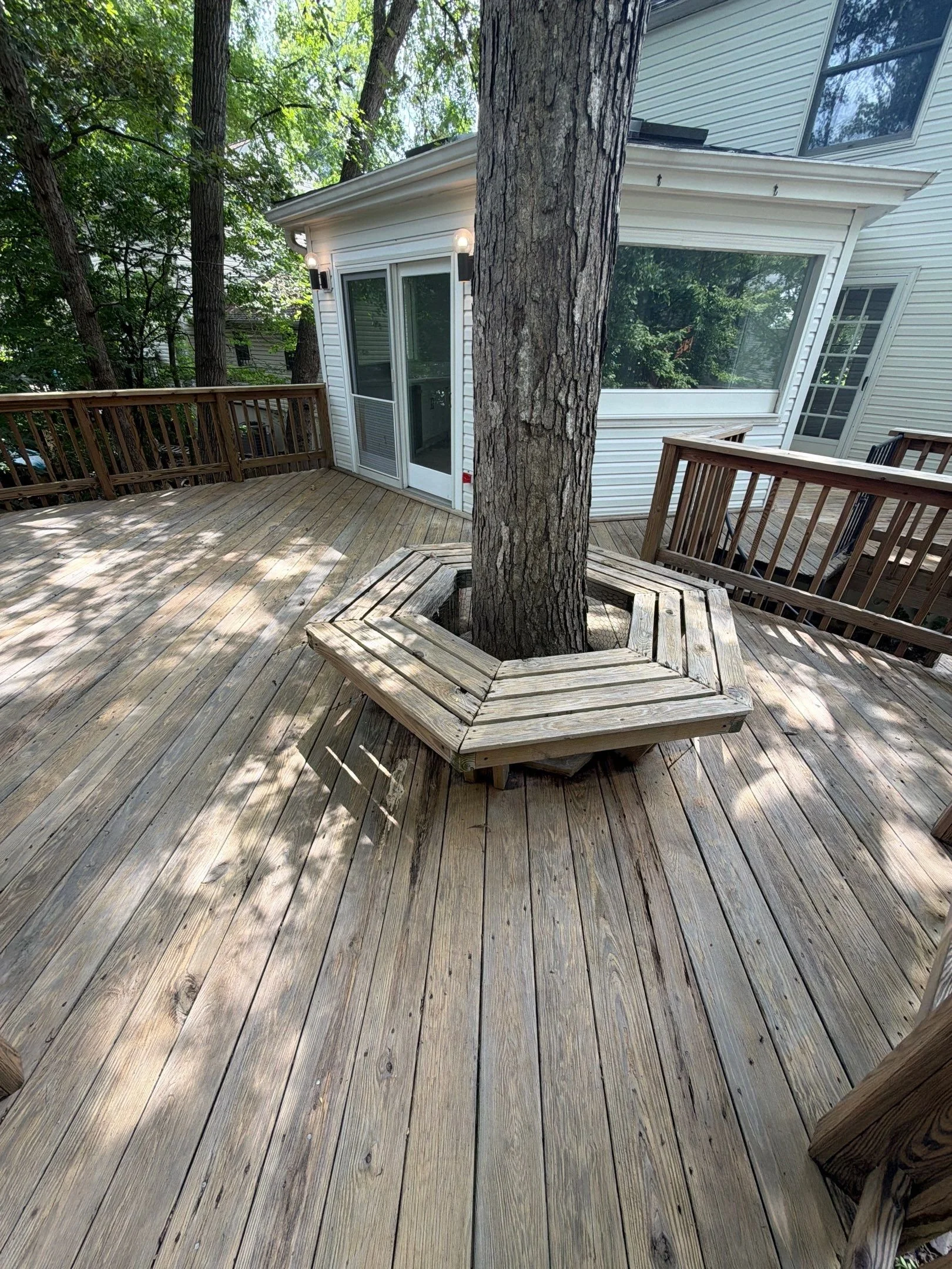 View of a wooden deck with trees and a house in the background, featuring a large tree in the center with a circular bench around it, and a white house with large windows.