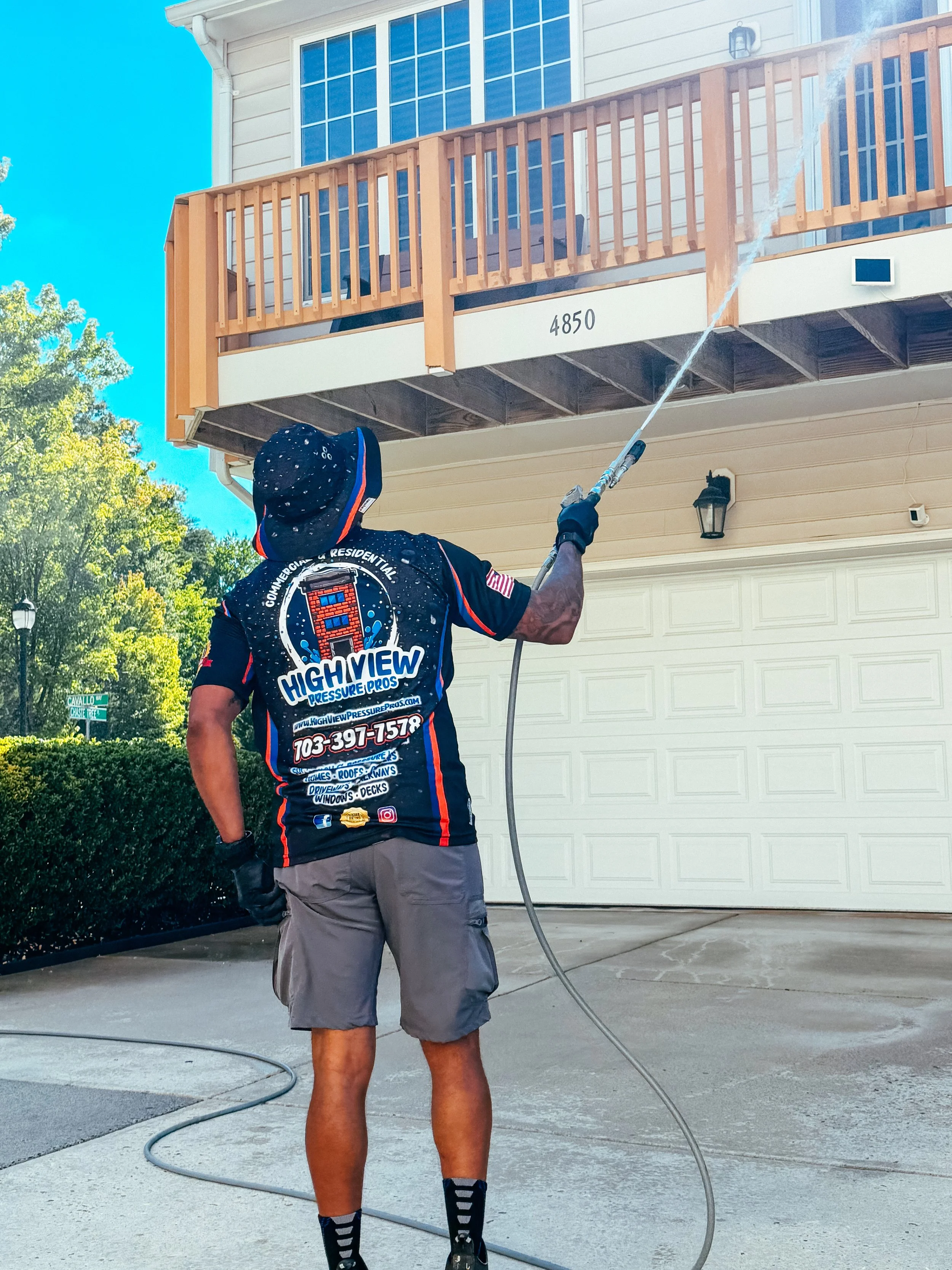 A person wearing a black hat, gray shorts, and a black shirt with colorful logos and text is pressure washing a house's second-story balcony railing and exterior wall on a sunny day. The person is standing on the driveway with trees and the house in the background.