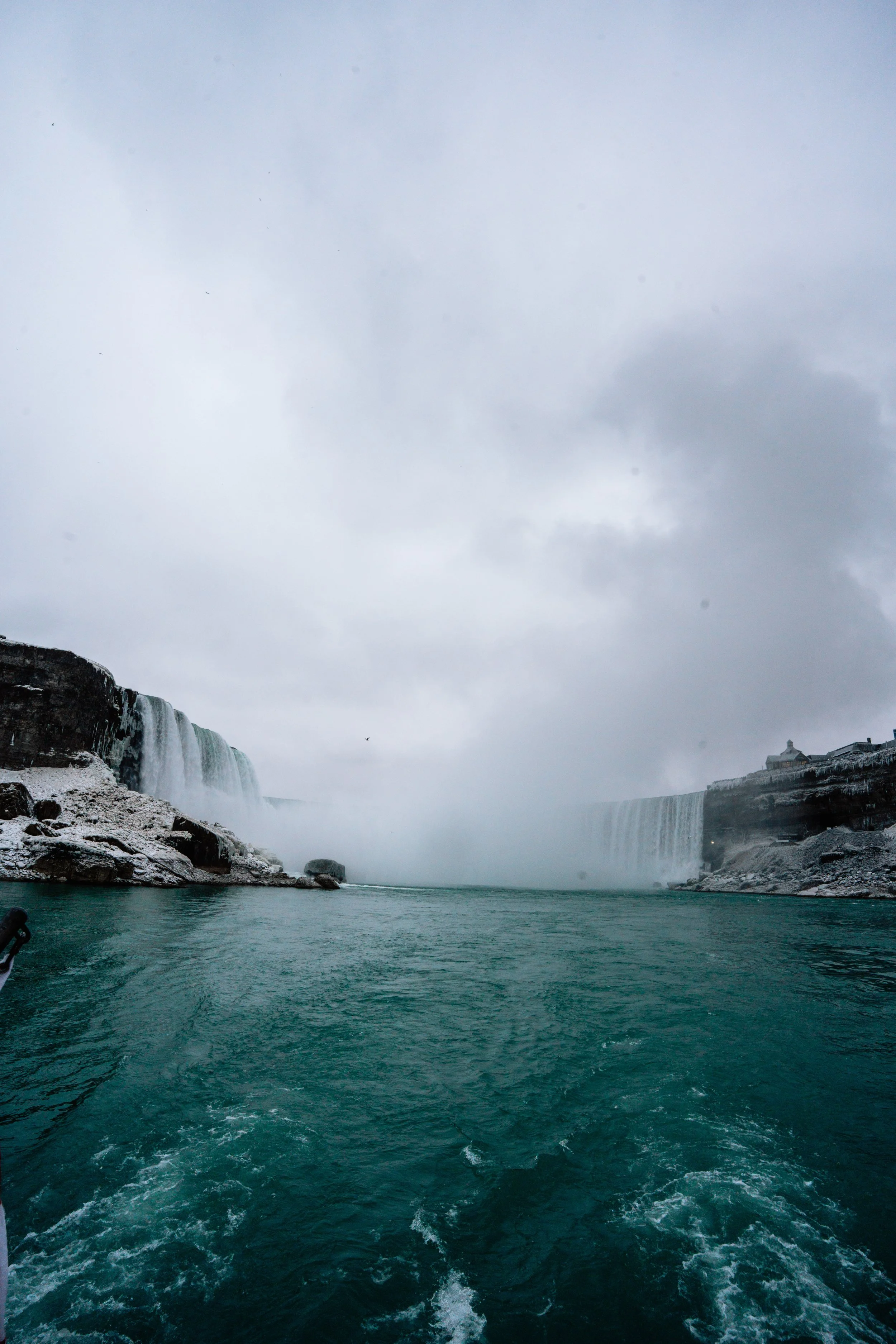 View of Niagara Falls with waterfalls on both sides, icy rocks, and cloudy sky in winter.
