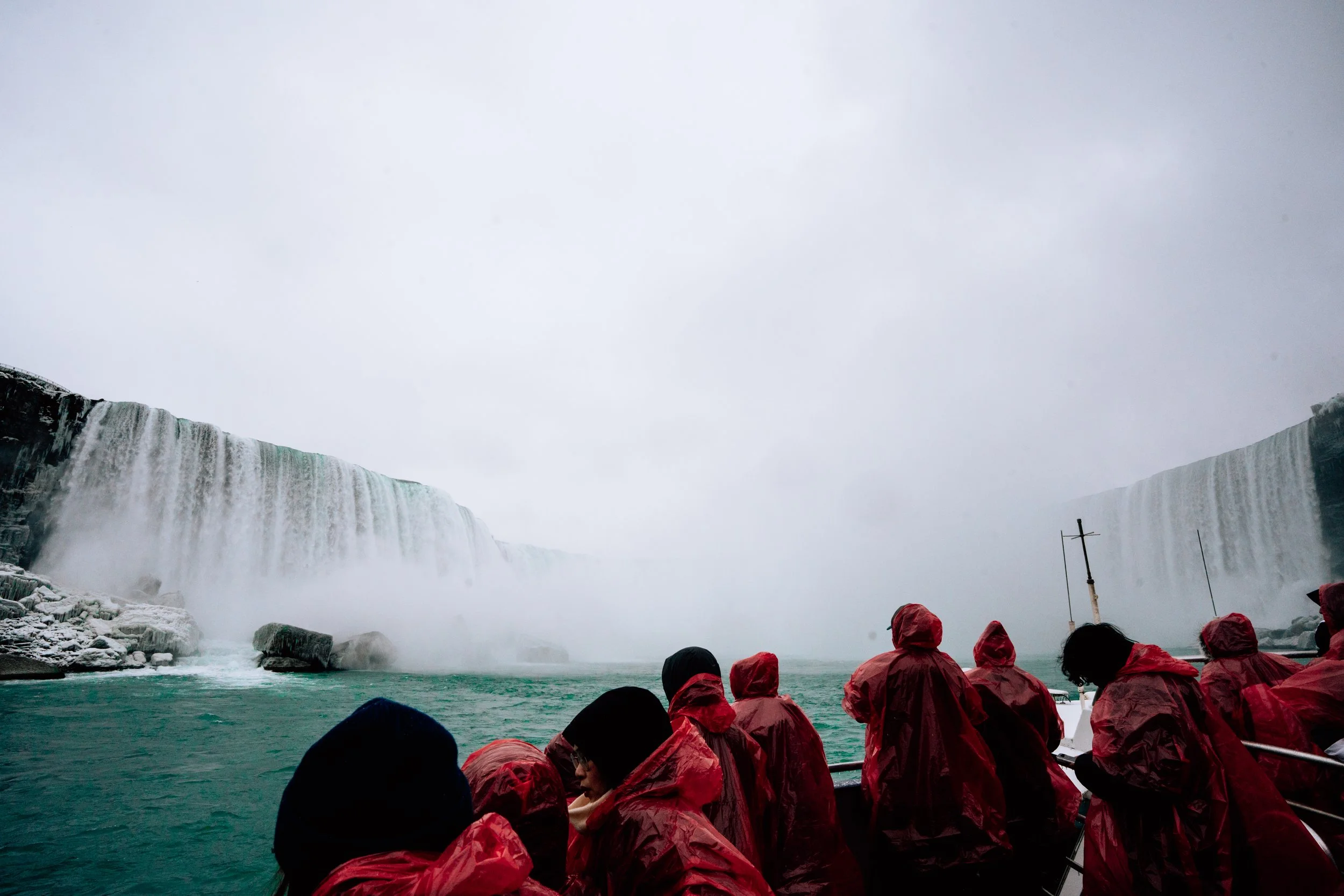 Tourists wearing red raincoats on a boat near Niagara Falls with mist rising from the waterfalls on a cloudy day.