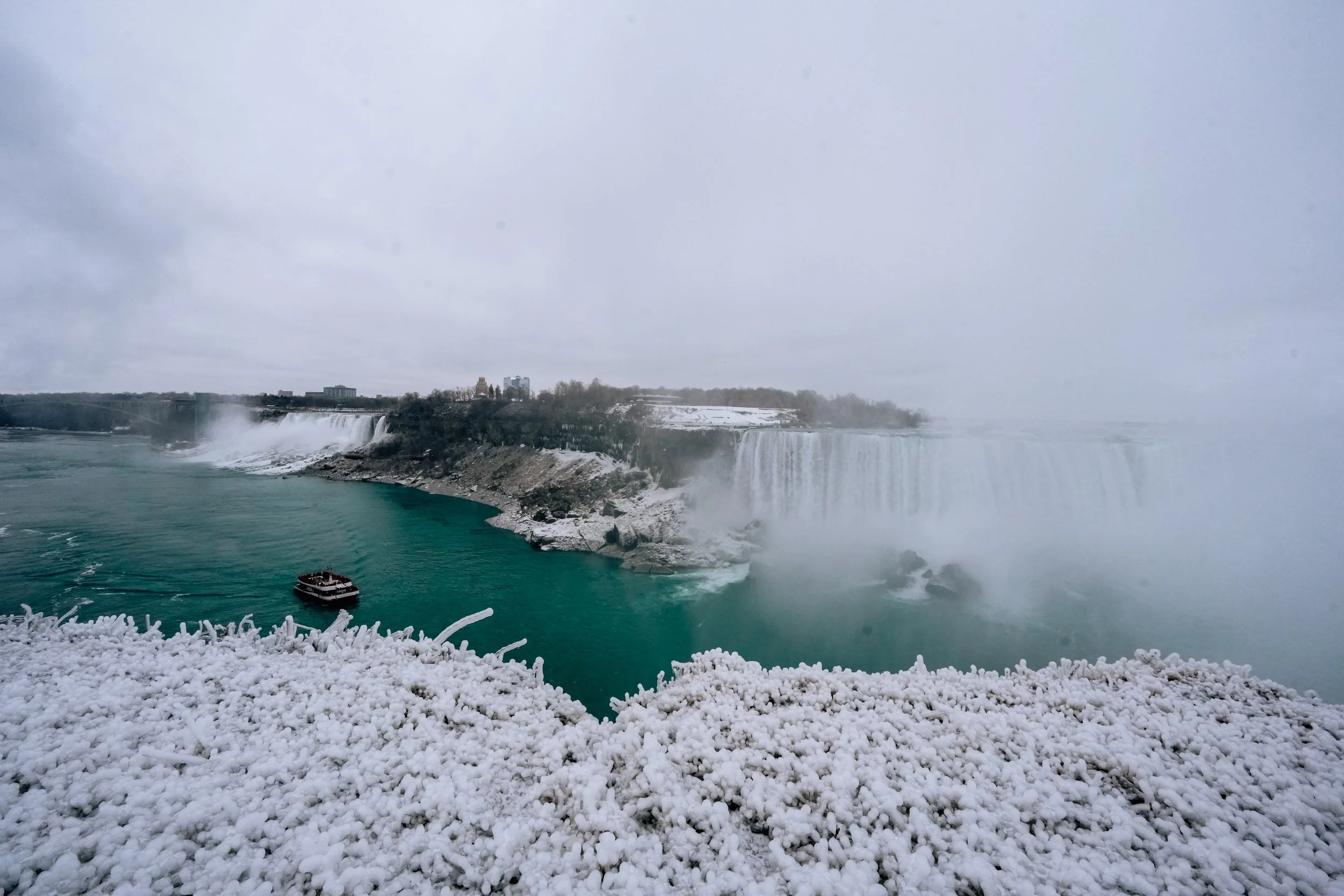 Niagara Falls partially covered in snow with a boat near the base of the falls on a cloudy day.