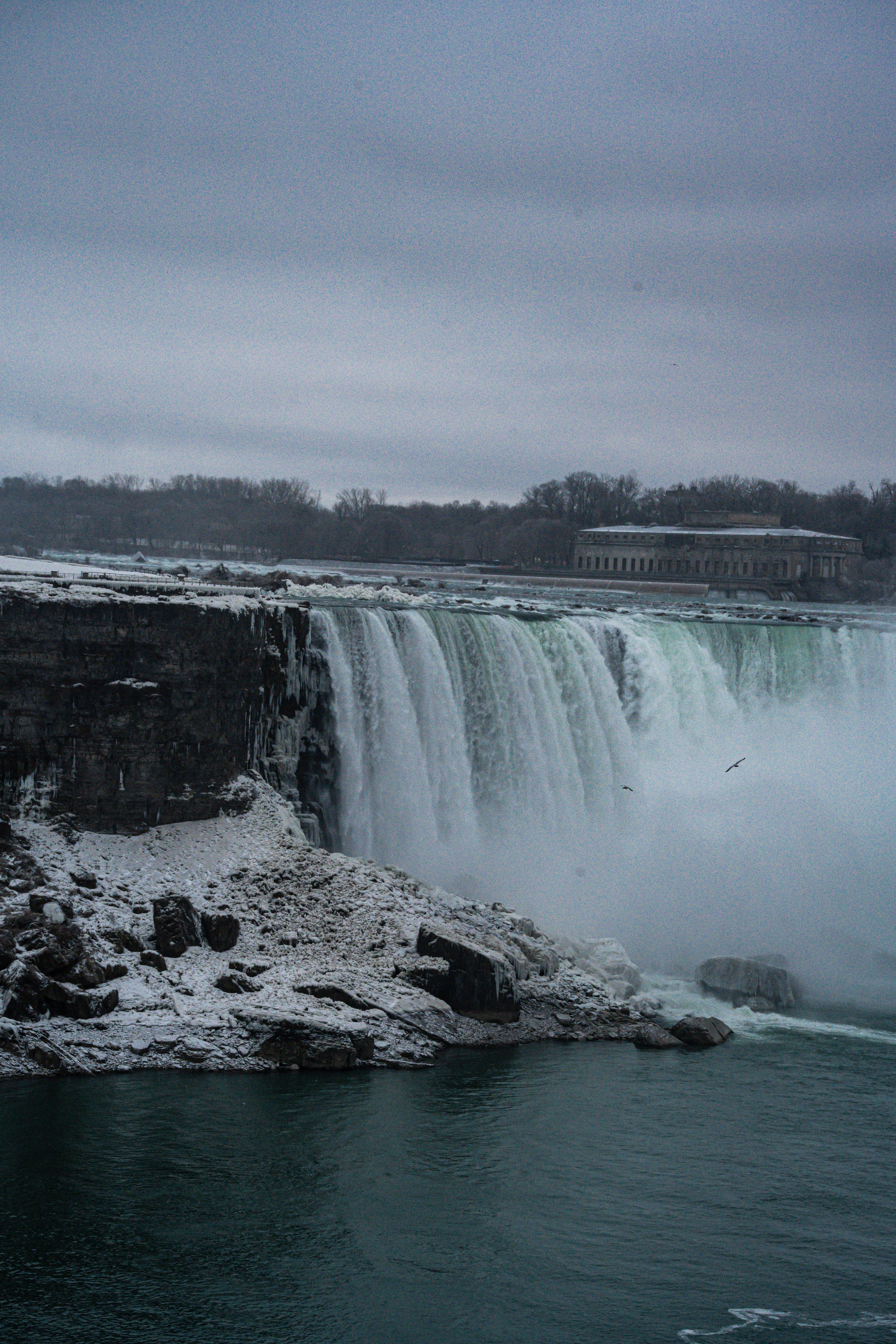 Niagara Falls during winter with partially frozen rocks and snow, cloudy sky, and mist rising from the waterfall.