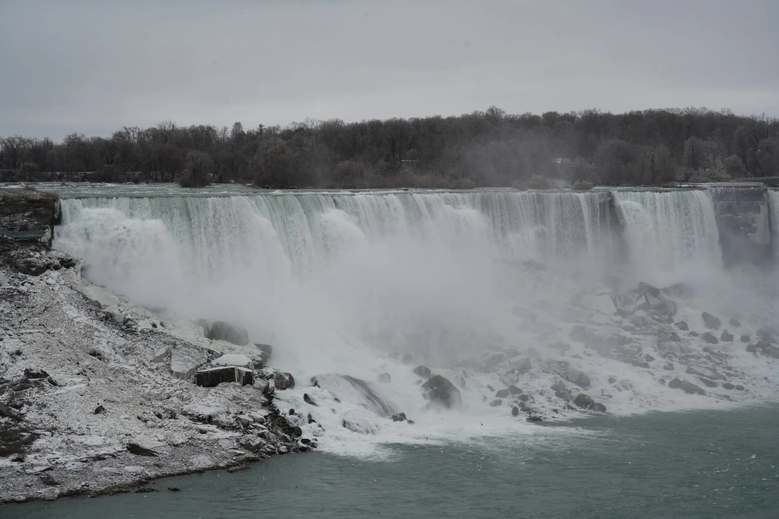A wide view of Niagara Falls with water rushing over the edge, surrounded by mist, rocks, and leafless trees under a cloudy sky.