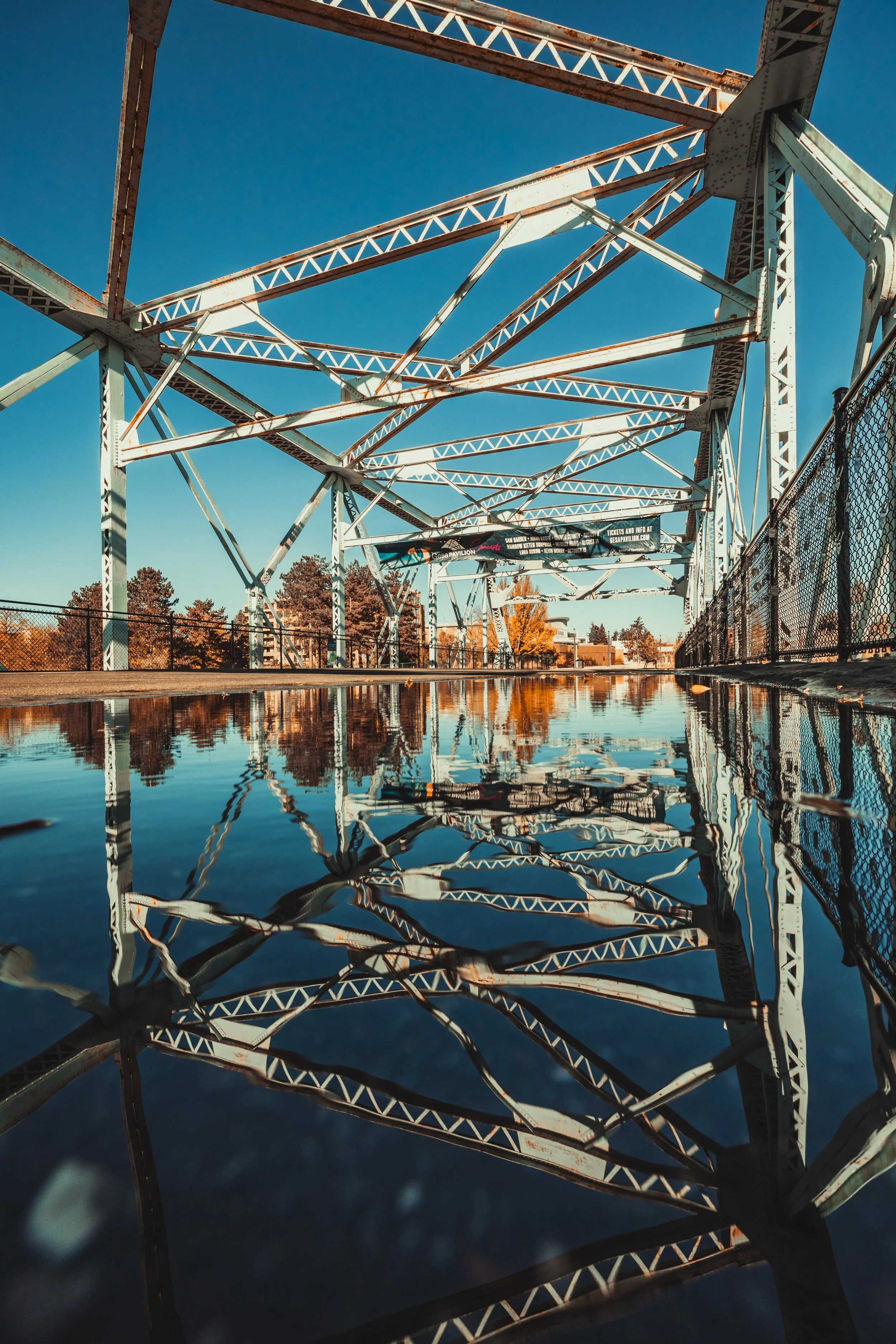 View of a metal bridge with a puddle below reflecting the structure, under a clear blue sky with some trees in the background.