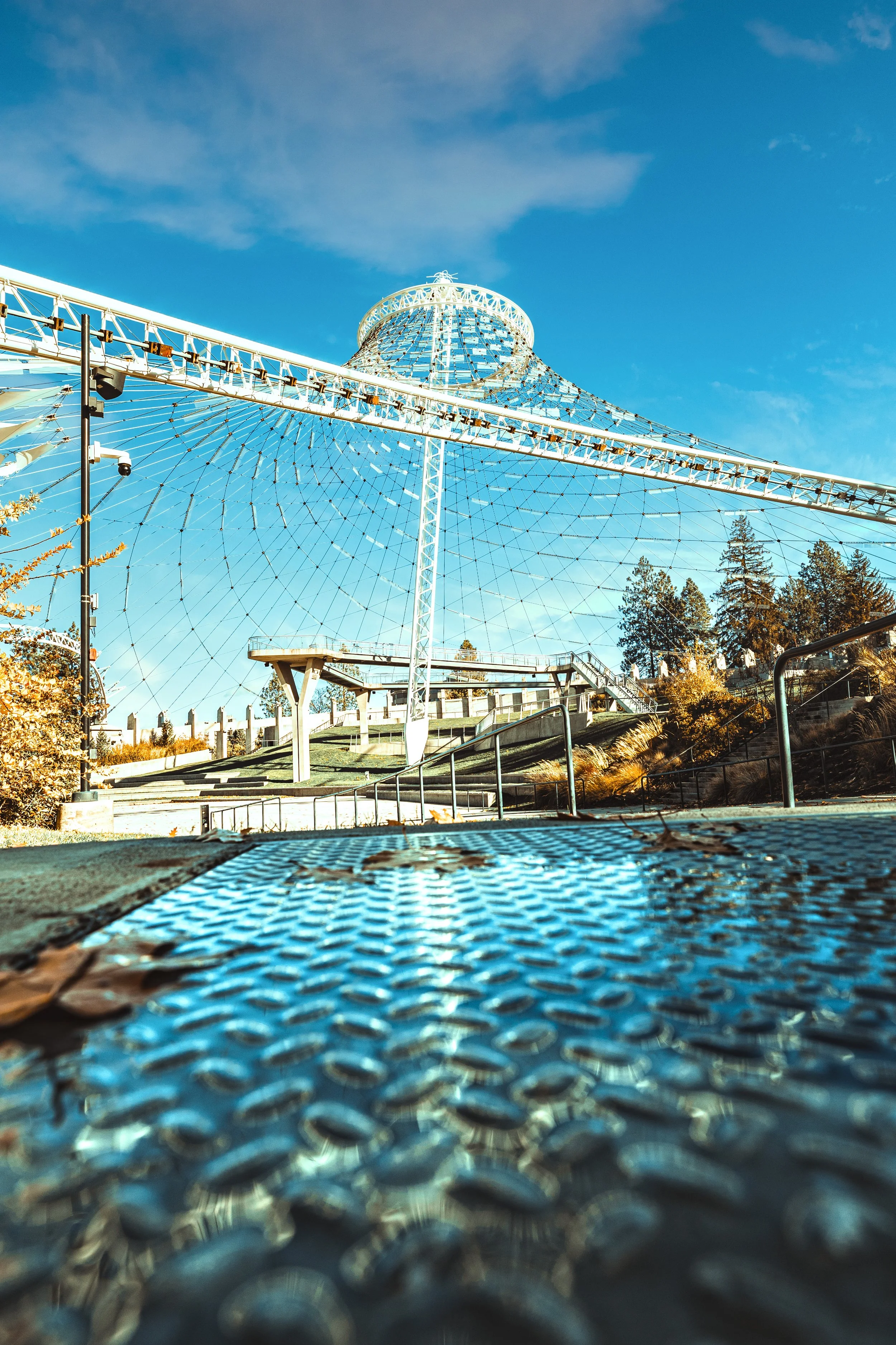 A large outdoor swing ride with a circular canopy and web-like structure underneath, set against a blue sky with some clouds, surrounded by trees and autumn foliage.