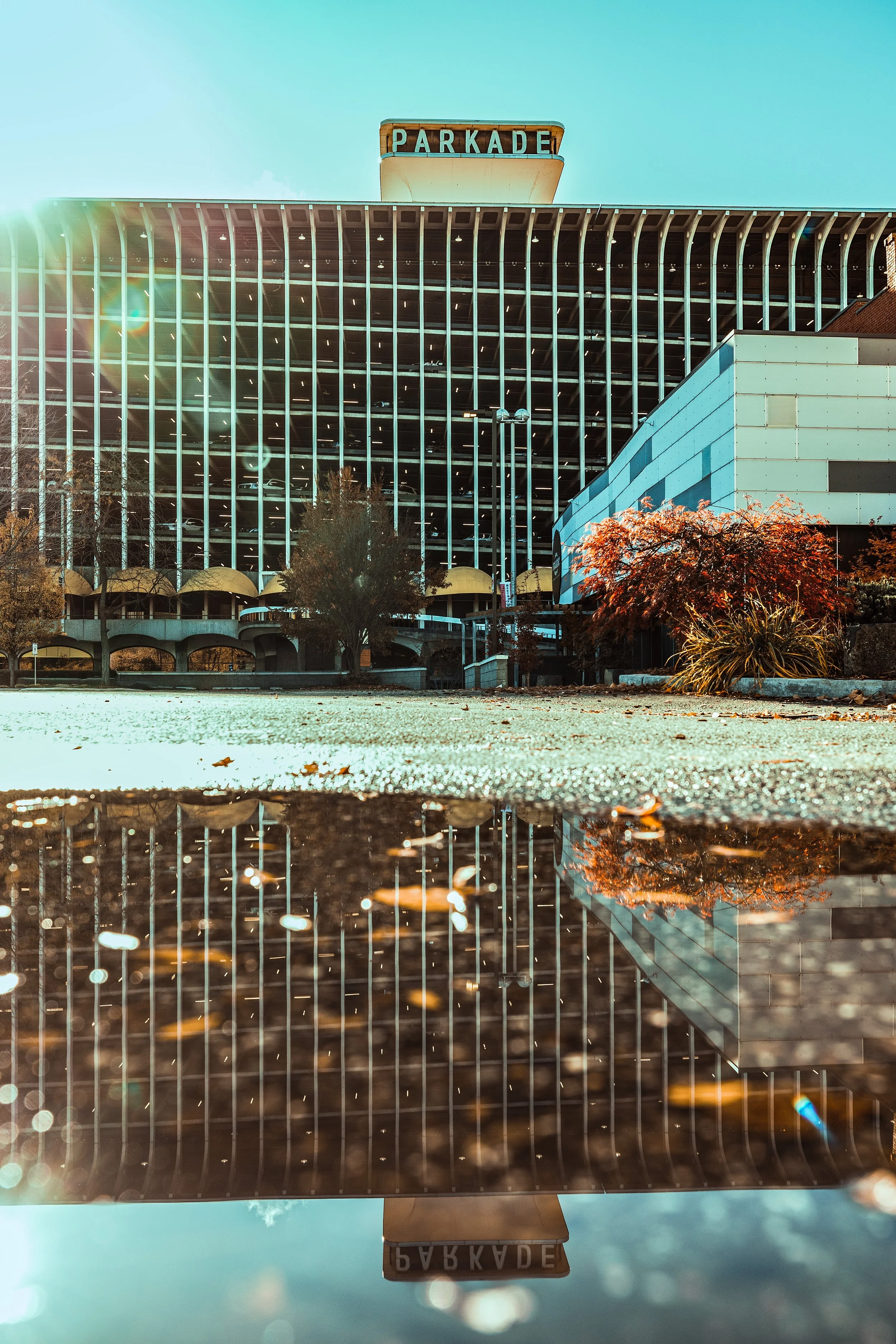 Front view of the Parkade parking structure with the sign at the top, reflected on a wet ground with fallen leaves and autumn trees in front of it.
