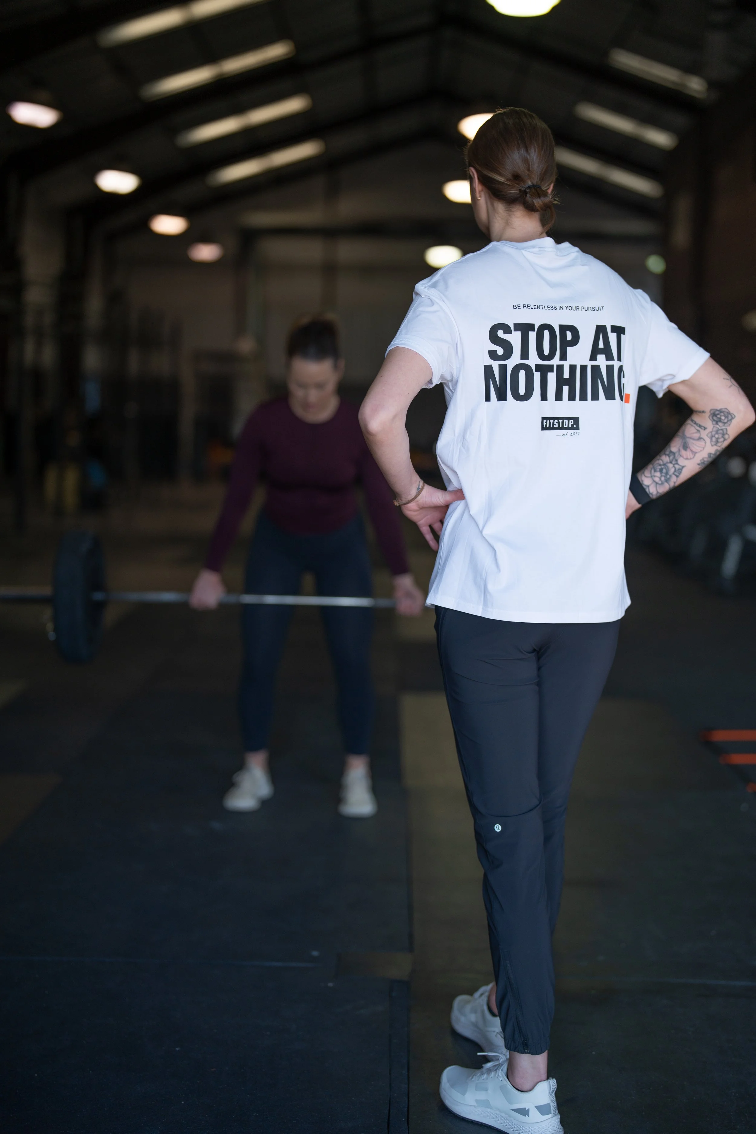 A woman with tattoos on her arm is observing another woman lifting a barbell in a gym. The woman watching is wearing a white T-shirt with the words 'STOP AT NOTHING' printed on the back.