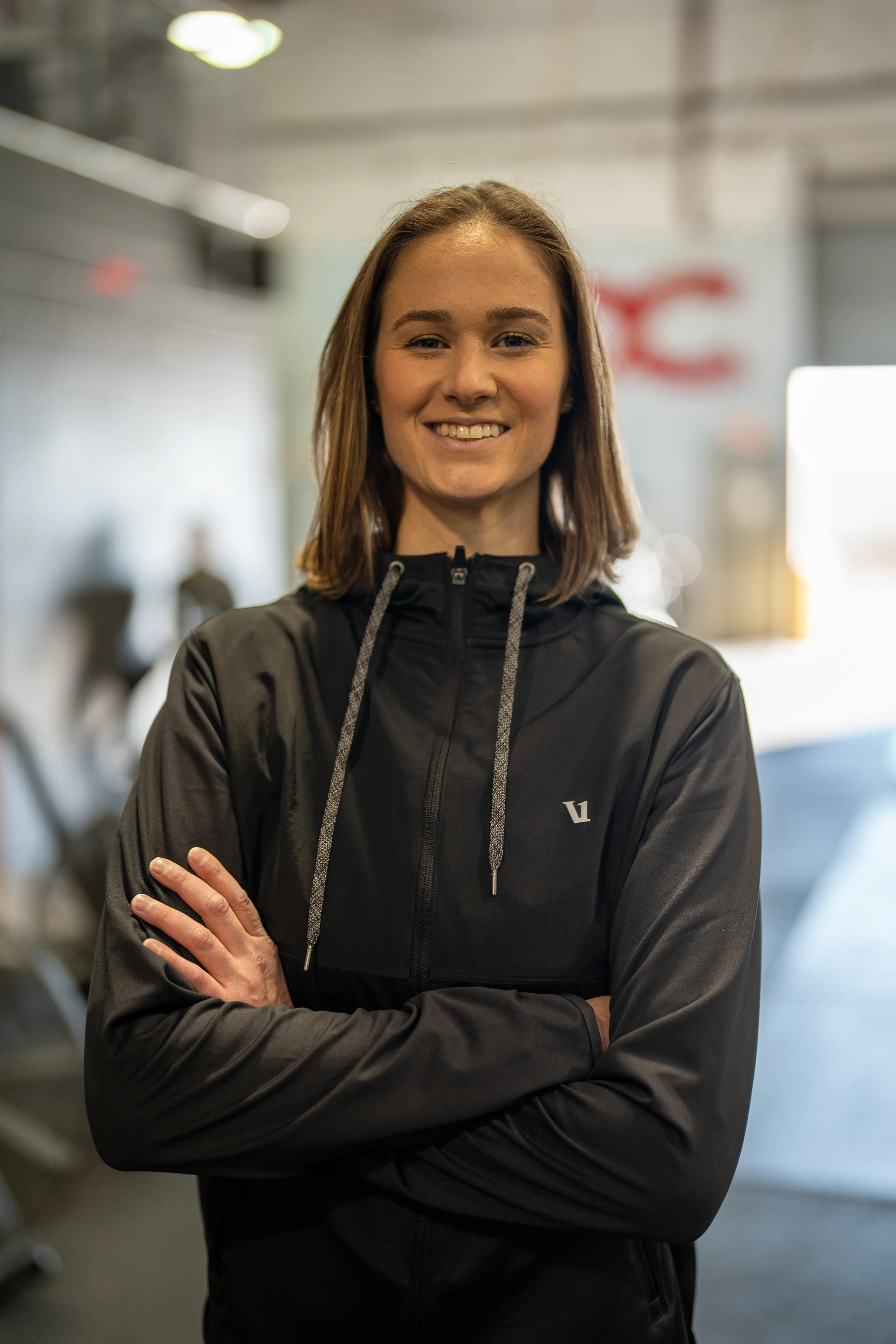 A young woman with shoulder-length brown hair, wearing a black athletic jacket with drawstrings, standing indoors with arms crossed, smiling at the camera.