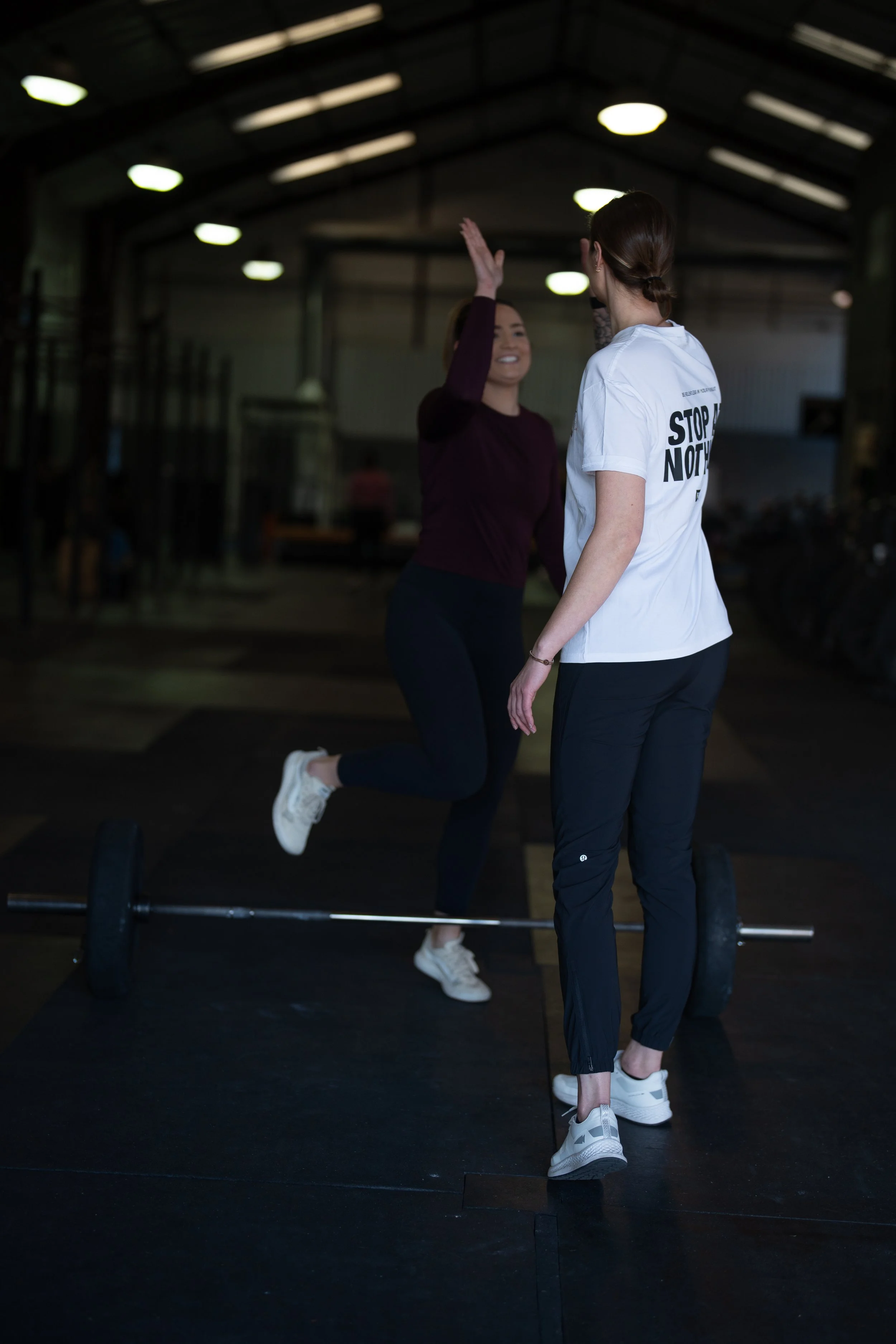 Two women in a gym, one performing a jump with a barbell on the floor, and the other coaching. The woman jumping is smiling, and the other woman wearing a white T-shirt with the message "Stop Not" turned away from the camera.