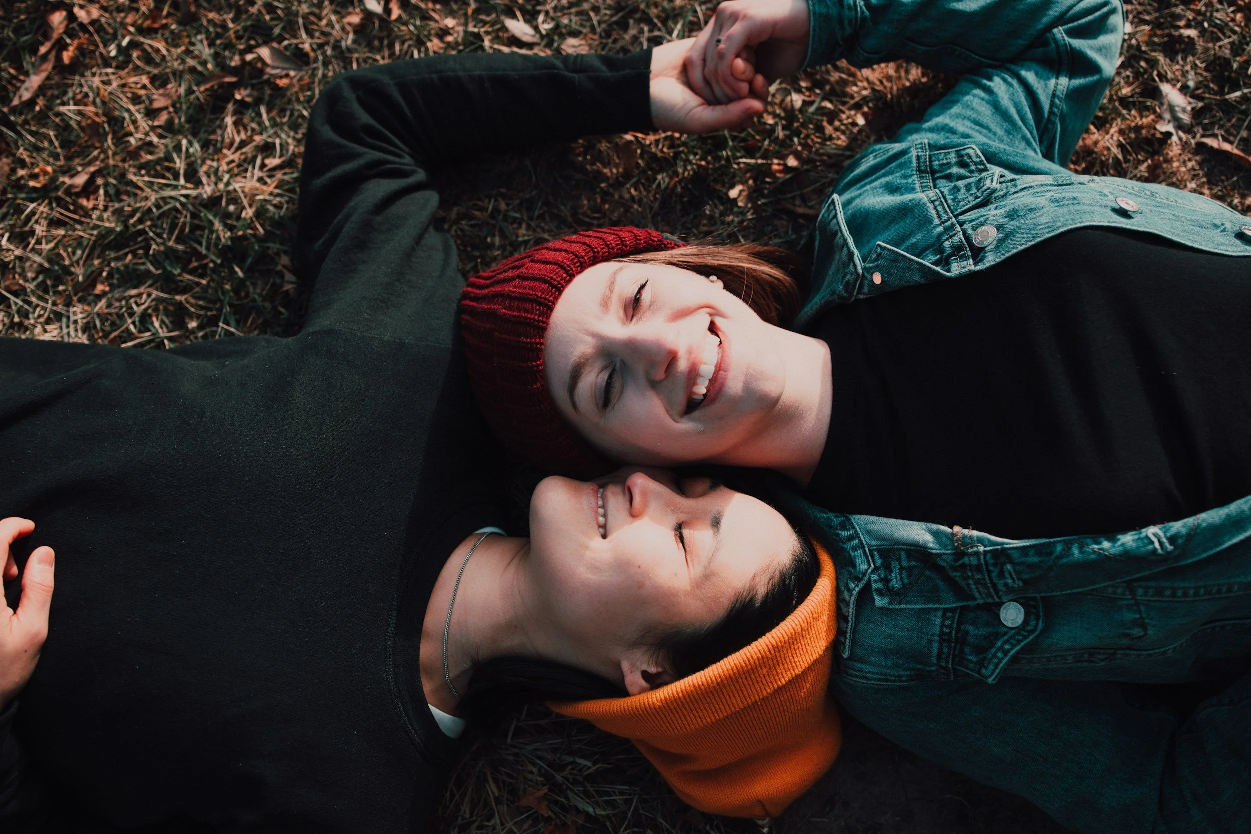 Two women lying on the grass holding hands and smiling, one wearing a black sweater and the other wearing a denim jacket and a red and orange knit hat.