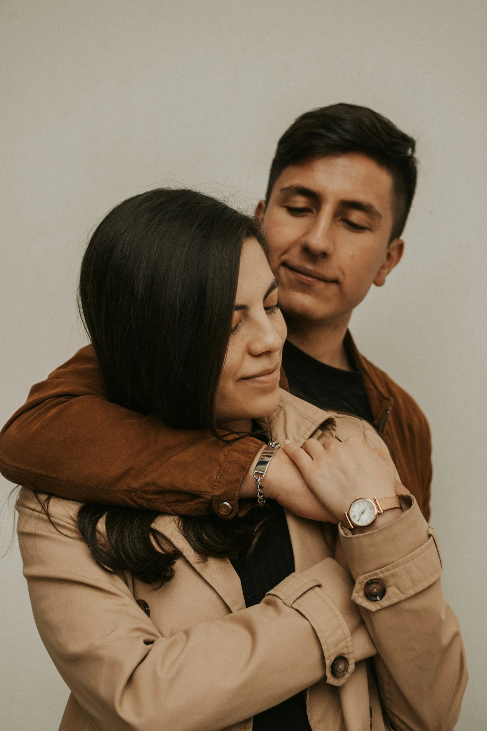 A man hugging a woman from behind, with both eyes closed and serene expressions, against a plain background.