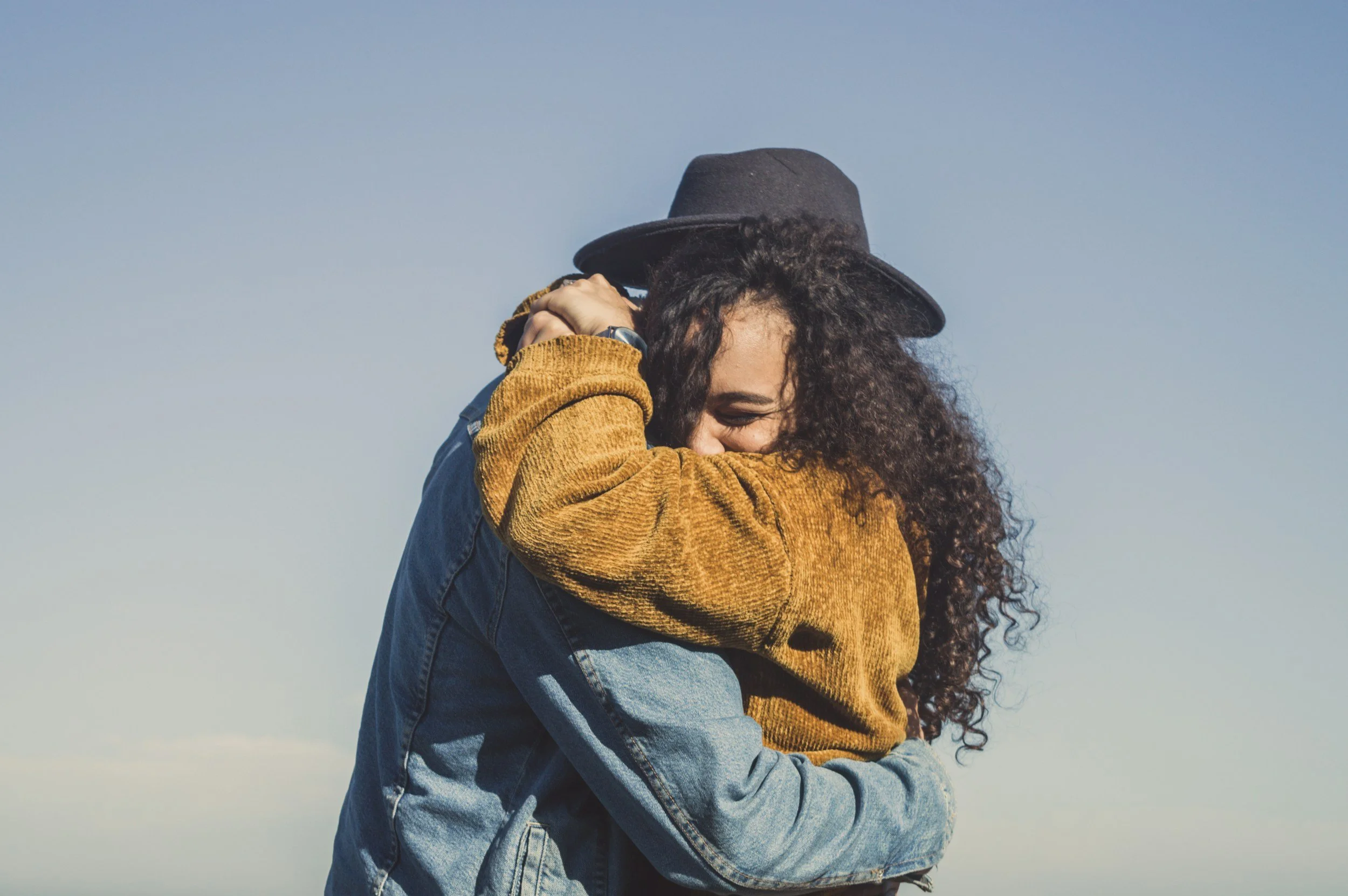 A woman with curly hair and a black hat hugging a person in a mustard yellow jacket and denim jacket outdoors against a clear blue sky