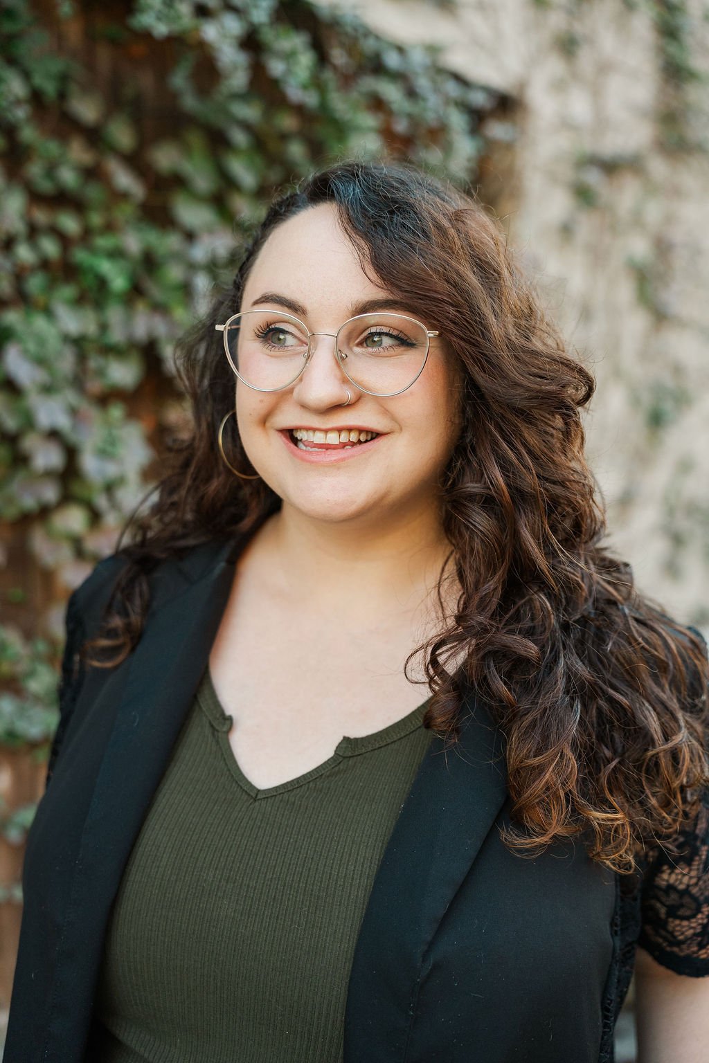 A woman with curly brown hair and glasses smiling outdoors, wearing a black blazer and green top, with a background of leaves and a stone wall.