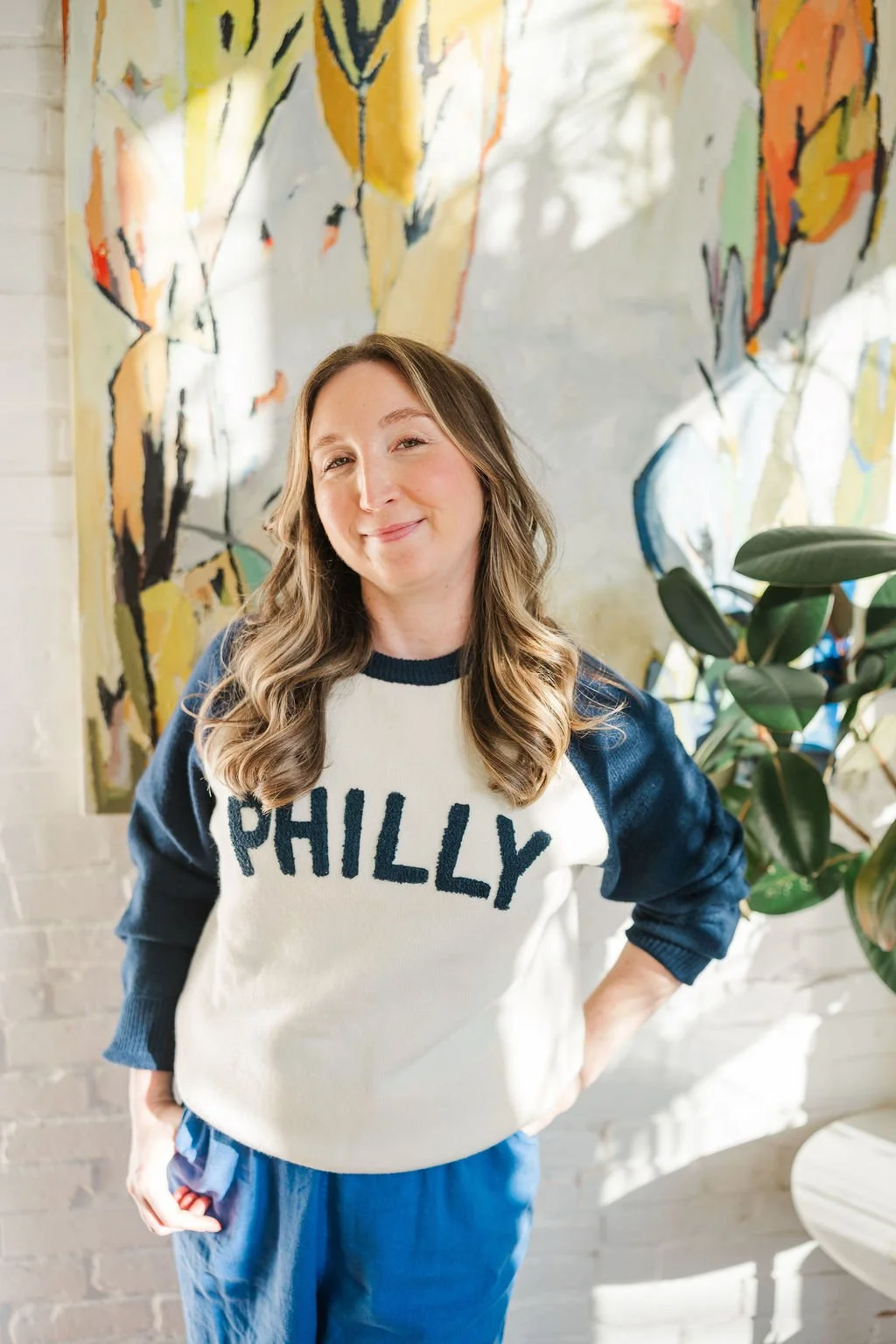 A woman with wavy brown hair smiling, wearing a white sweatshirt with dark blue sleeves and 'PHILLY' written on it, standing in front of an abstract colorful painting and a green plant.