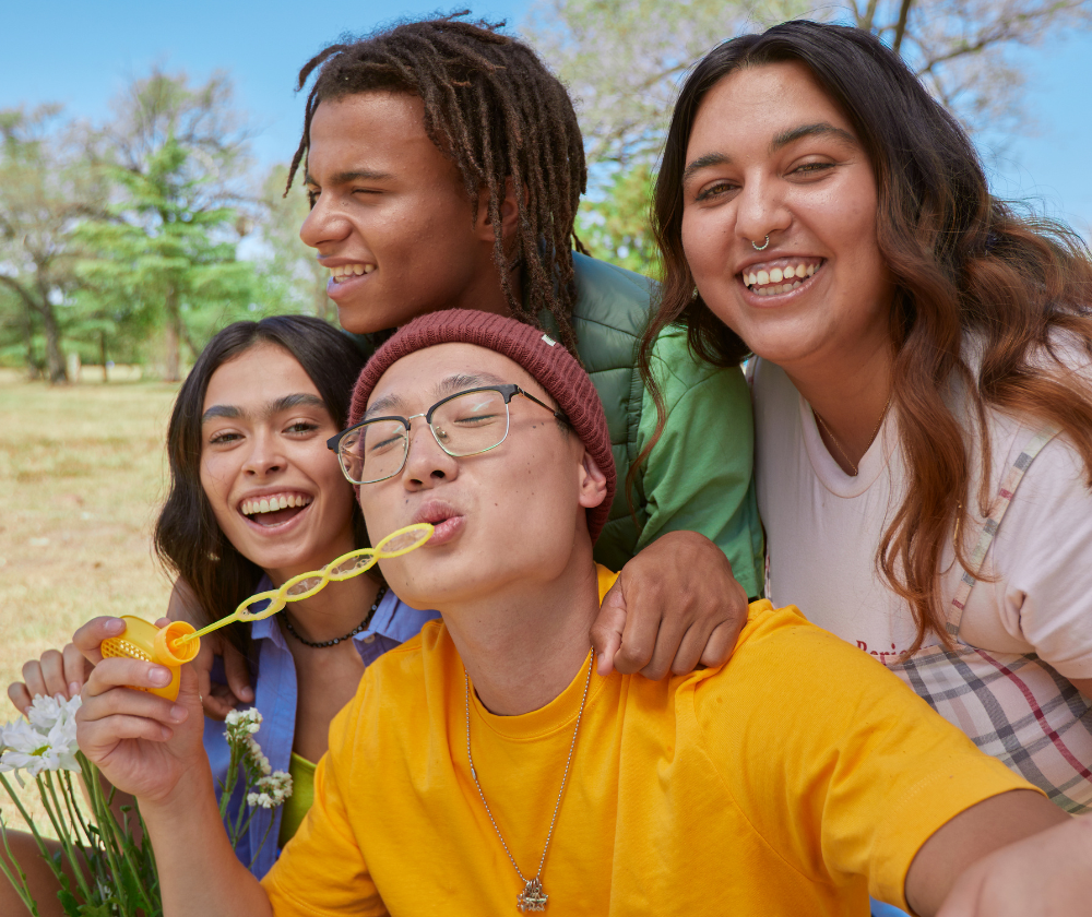 Four young friends smiling and posing for a selfie outdoors with trees and blue sky in background.