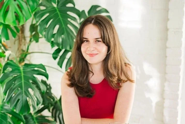 A young woman with long, wavy brown hair smiling and crossing her arms, wearing a red sleeveless top, standing indoors with a colorful artwork and a window in the background.